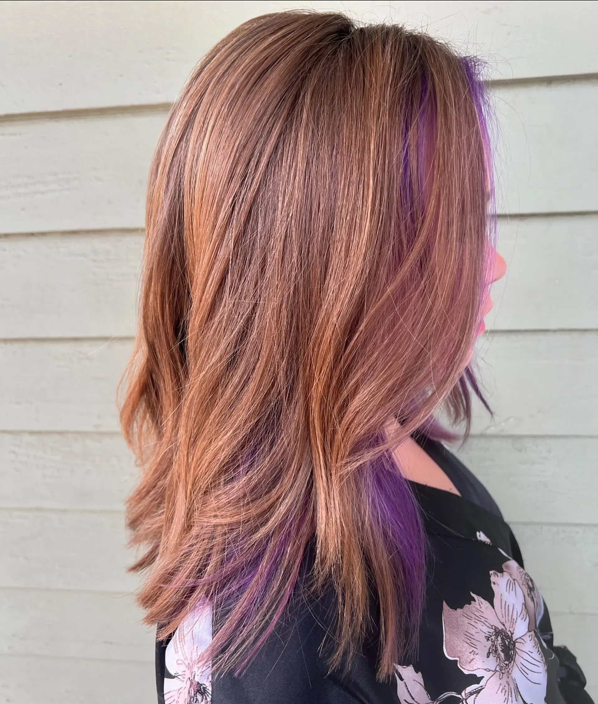 A medium-length, layered cut featuring soft waves. The hair is colored with a dimensional rose gold and warm brown base, highlighted by striking purple peek-a-boo sections underneath. 1