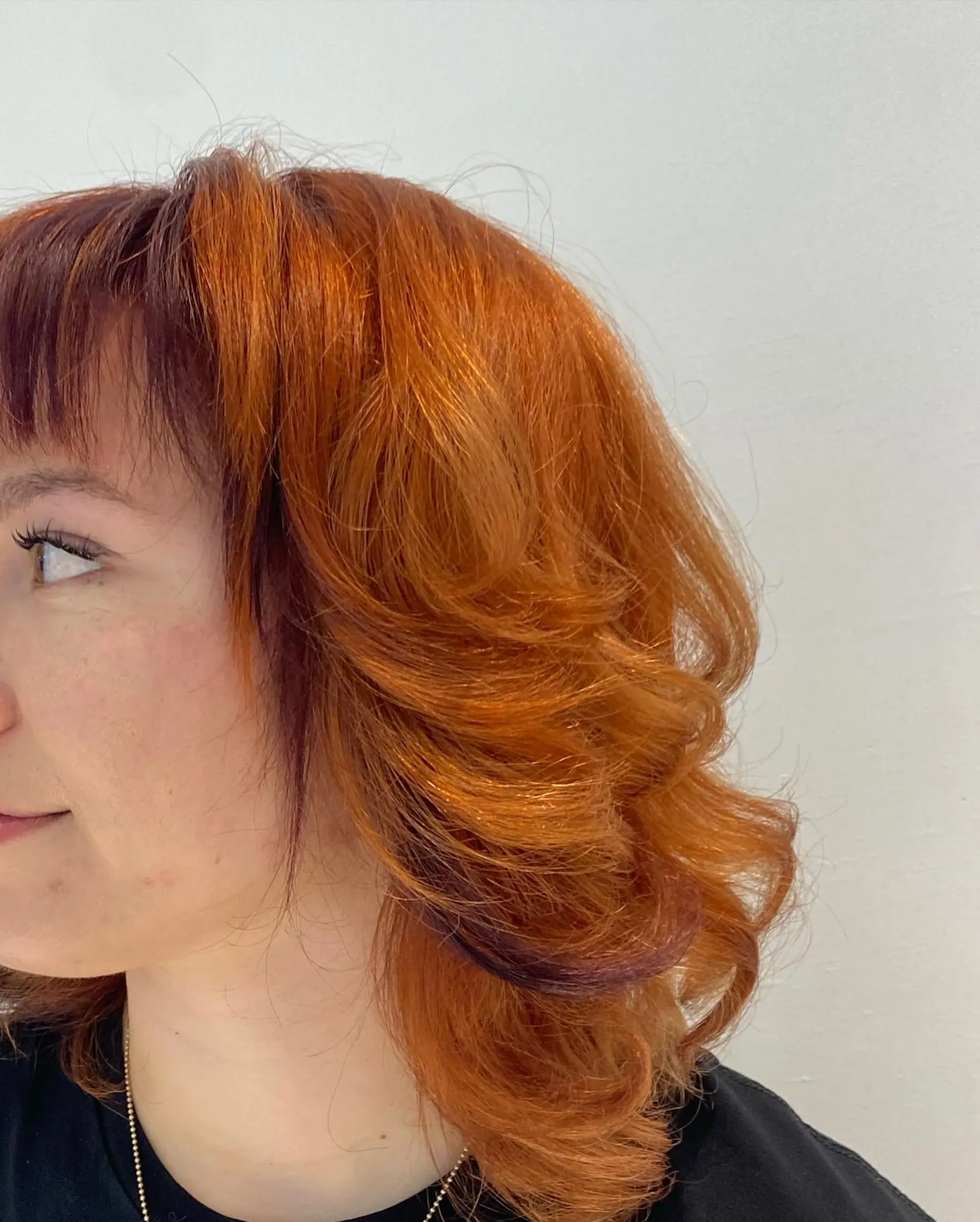 Medium-length layered copper hair styled in bouncy voluminous curls with blunt textured micro-bangs and subtle dark undertones. 1
