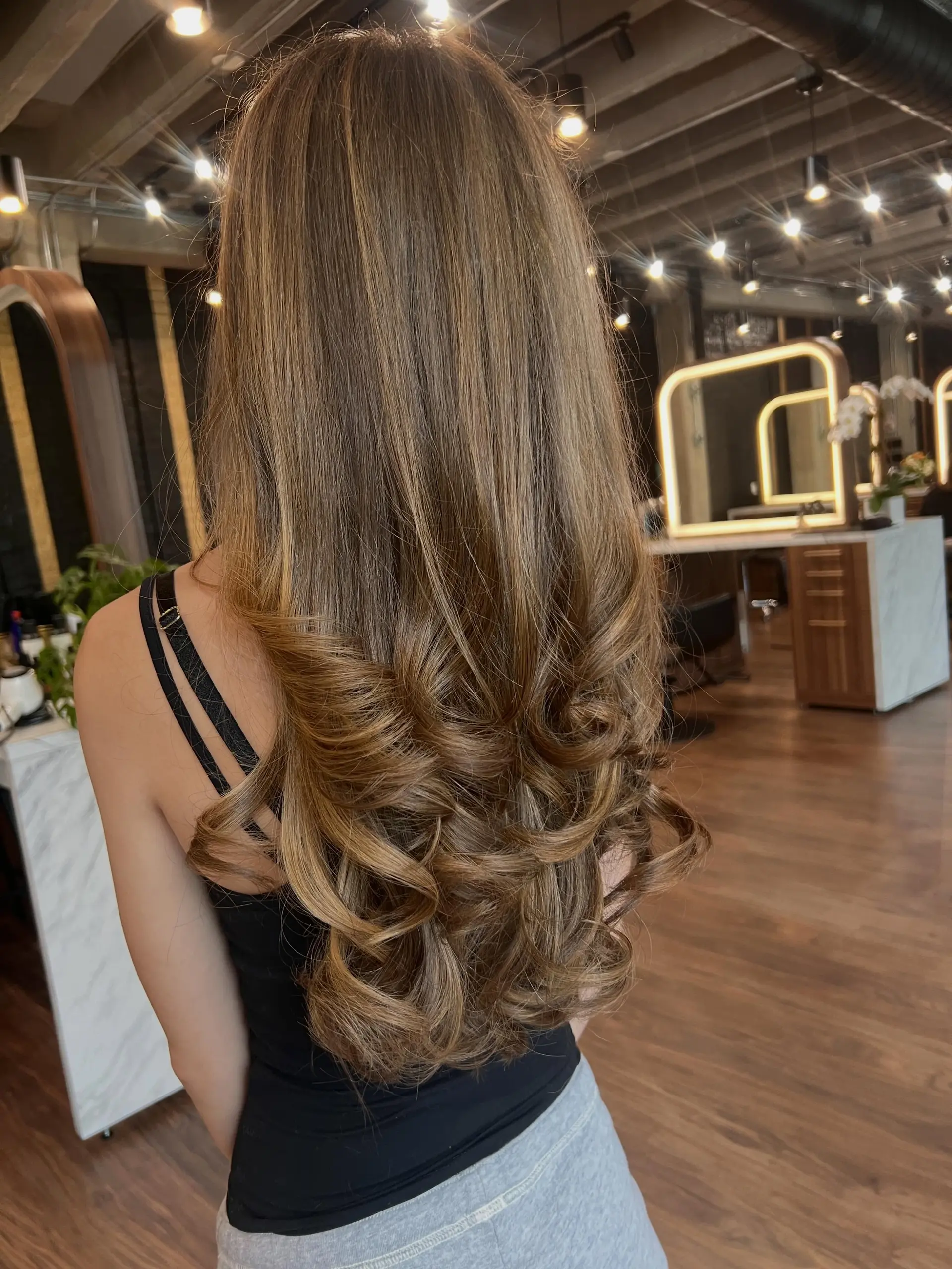 Long layered bronde hair worn smooth through the crown and mid-lengths, transitioning into polished, voluminous curls at the ends. 1