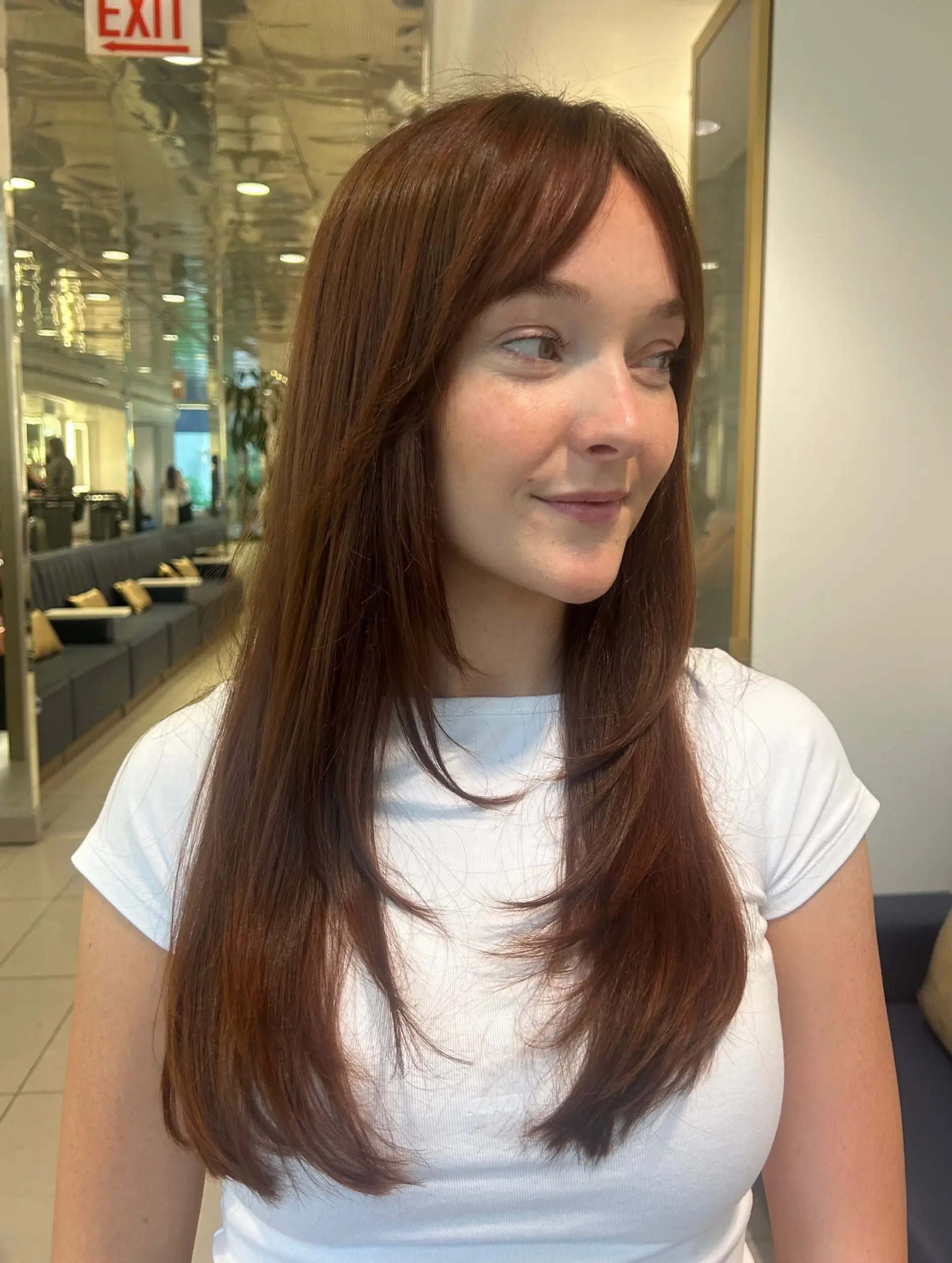 Long auburn hair cut with soft, graduated face-framing layers and parted curtain bangs, styled smooth and straight. 1