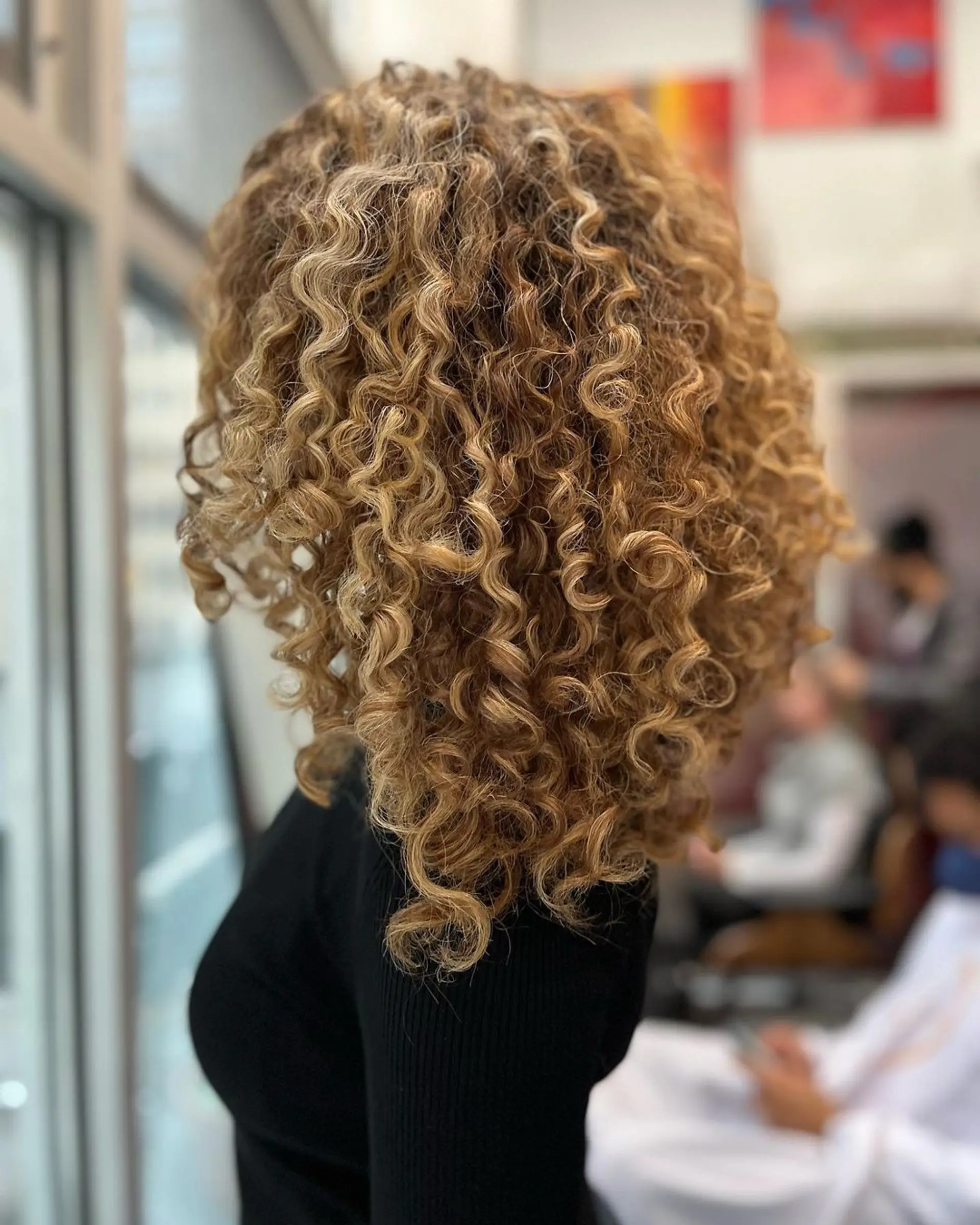 Medium-length curly hairstyle with honey blonde and golden brown hues. Natural curls form bouncy, voluminous ringlets with highlights, framing the face beautifully. 3