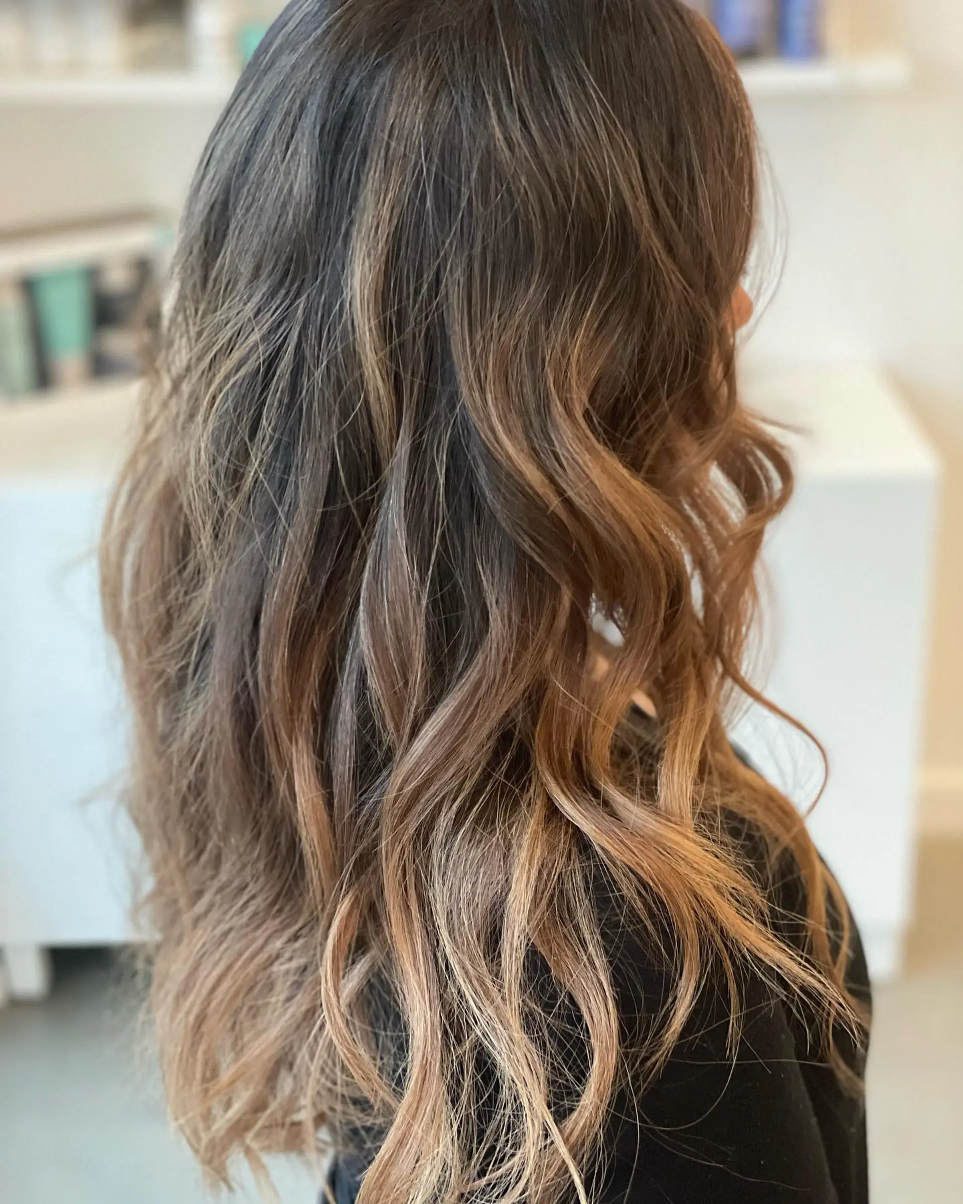 Long wavy hairstyle featuring a deep brunette base seamlessly blending into warm honey bronde balayage ribbons. 1