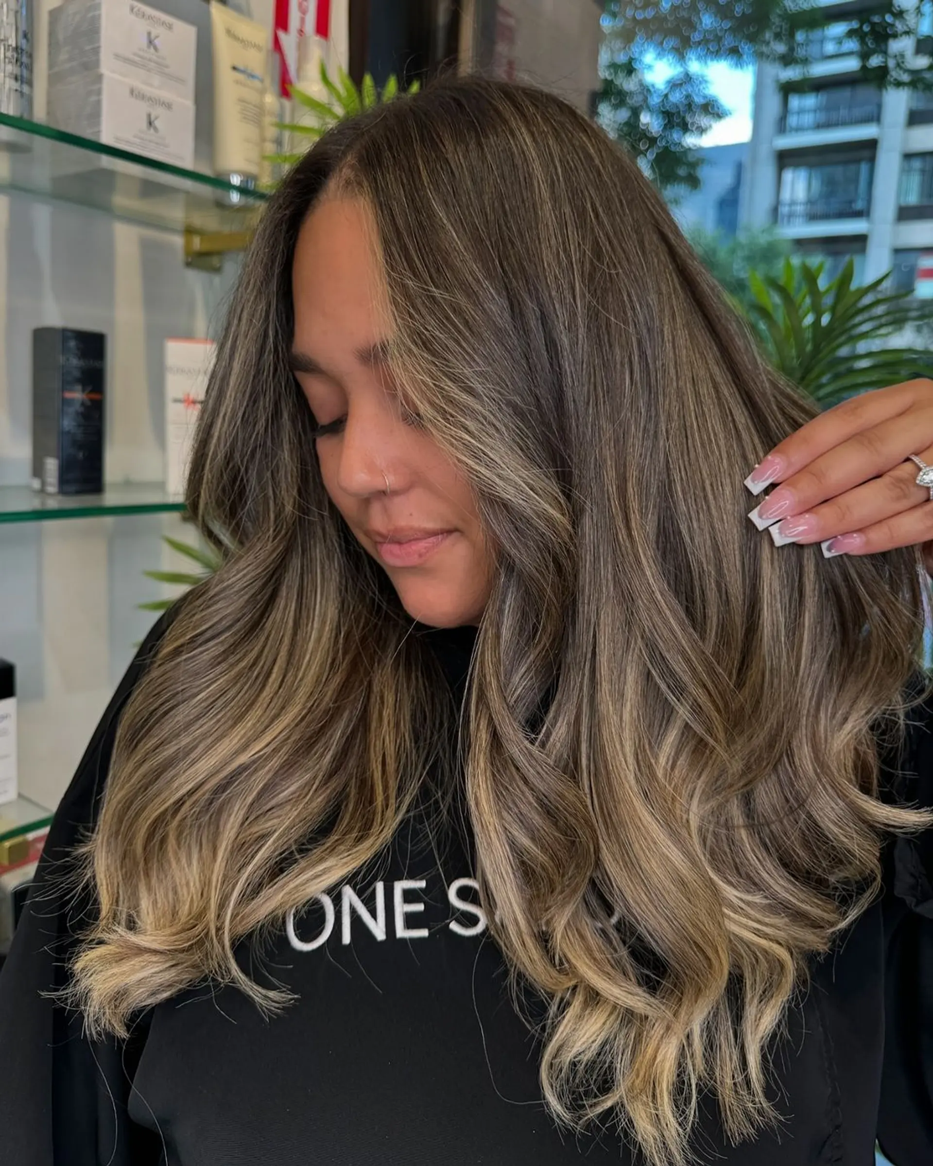 This long, wavy hairstyle features dark brown roots transitioning into light brown and honey blonde through balayage. The soft, beachy waves are complemented by layered cuts and face-framing highlights, adding movement and dimension. 1
