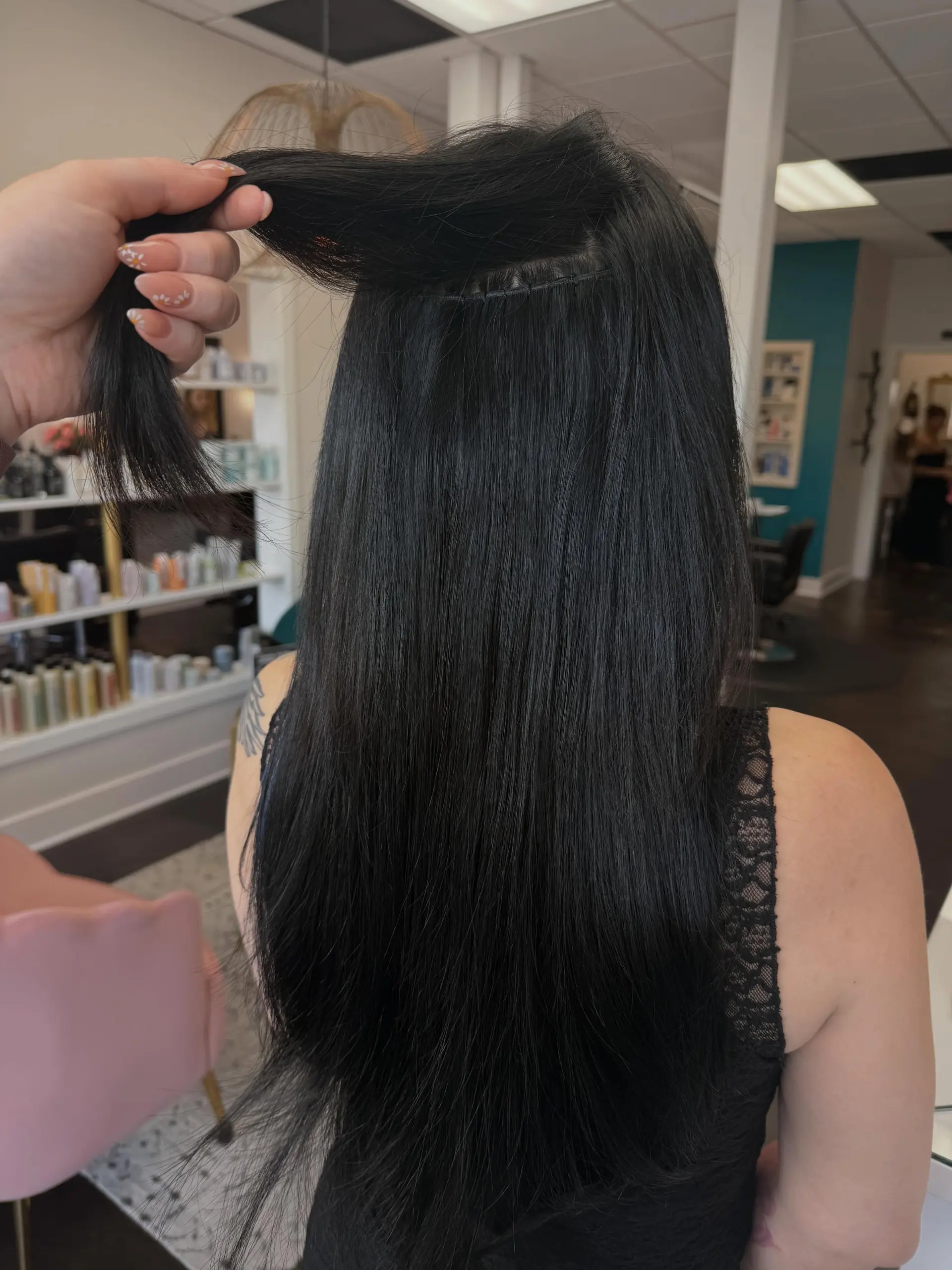 A client displays long, straight jet-black hair as the stylist lifts a section to showcase the seamless hand-tied extension installation underneath. 1