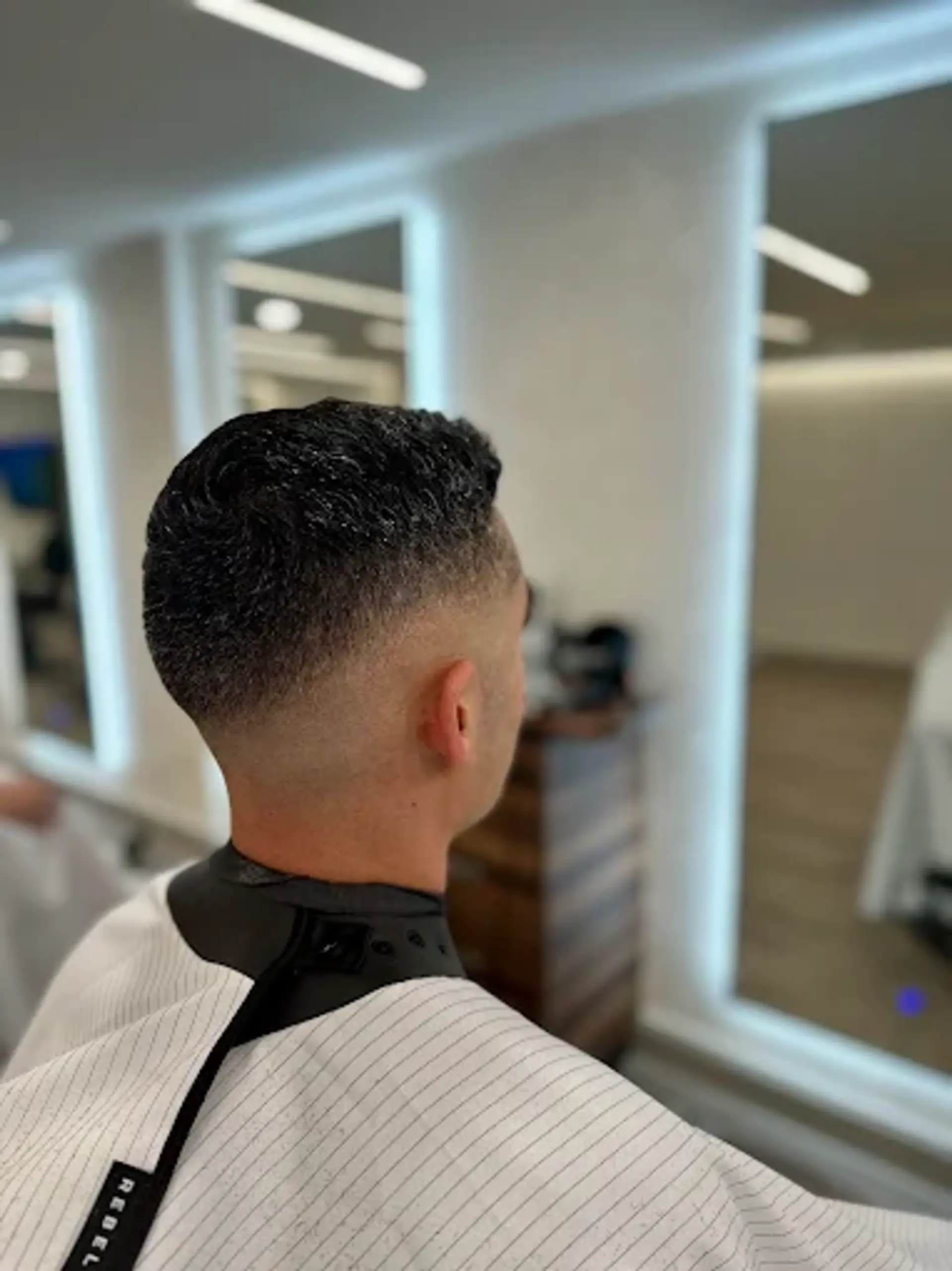 A sharp high skin fade where the sides and back are clipped very short, almost to the skin, and gradually fade up to a short, wavy, and textured top. The top hair is styled forward and slightly up, showcasing its natural waves and adding volume. 1