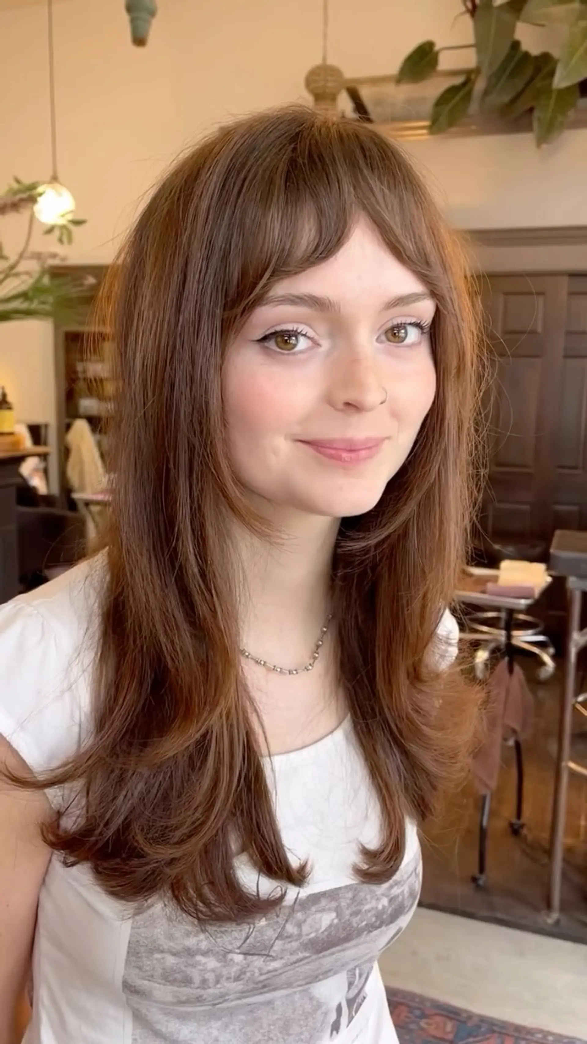 Long warm brown haircut featuring soft cascading layers, face-framing pieces, and arched baby bangs. 1