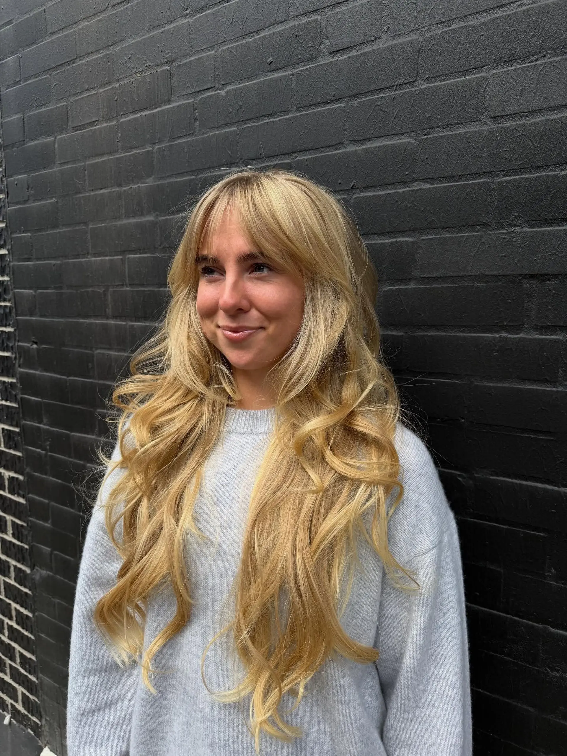 Very long warm blonde hair styled with soft, cascading loose waves, face-framing layers, and textured curtain bangs. 1