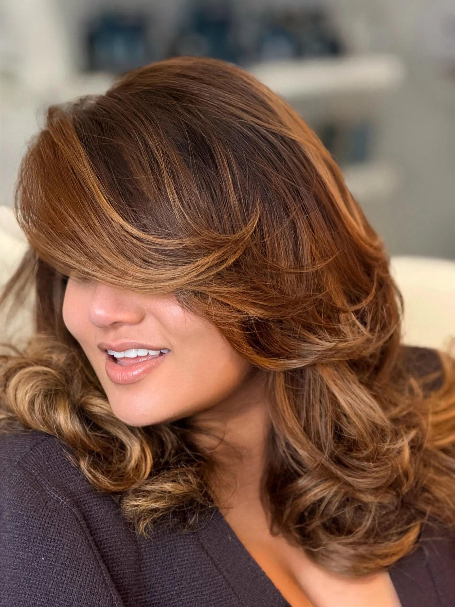 Voluminous layered blowout featuring swooping side-swept bangs and soft waves in warm caramel-brown tones. 1