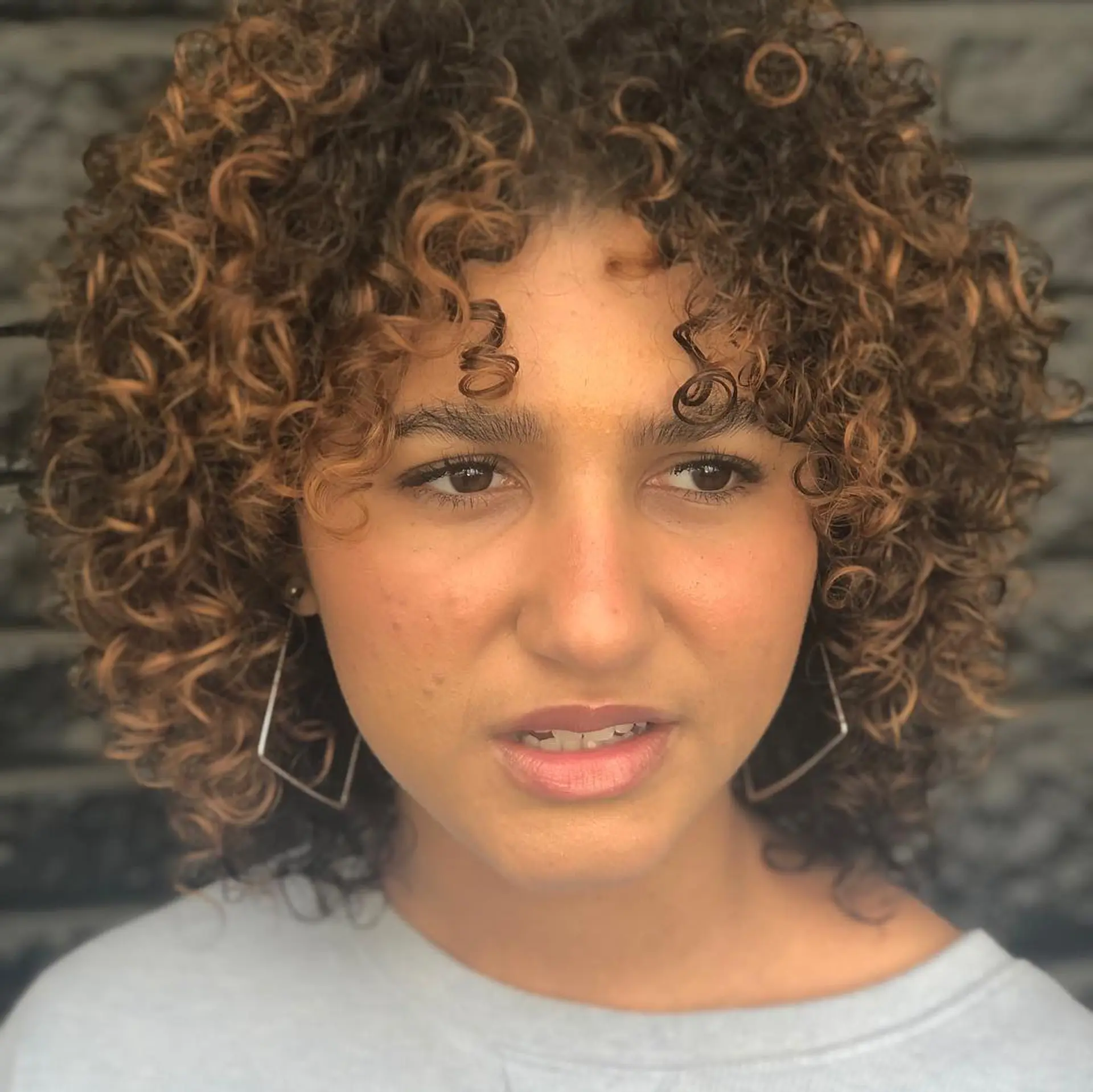 A layered, shoulder-length curly haircut featuring defined ringlets, natural volume, and face-framing curly bangs with warm caramel highlights. 1