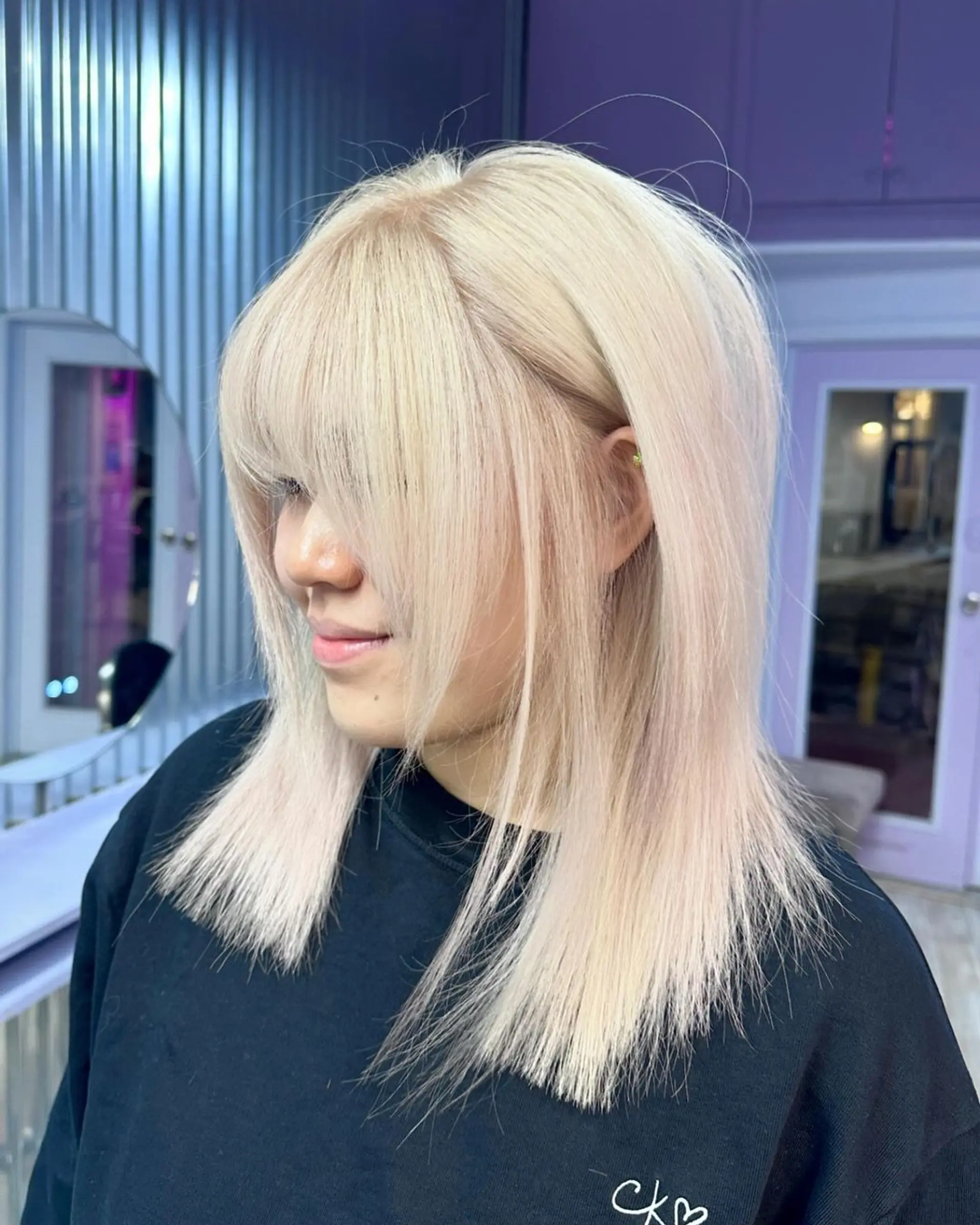A sleek, medium-length platinum blonde hairstyle with full, straight-across bangs and face-framing layers. The straight hair has a soft, shiny finish and a light, airy texture, creating a modern and chic look. 2