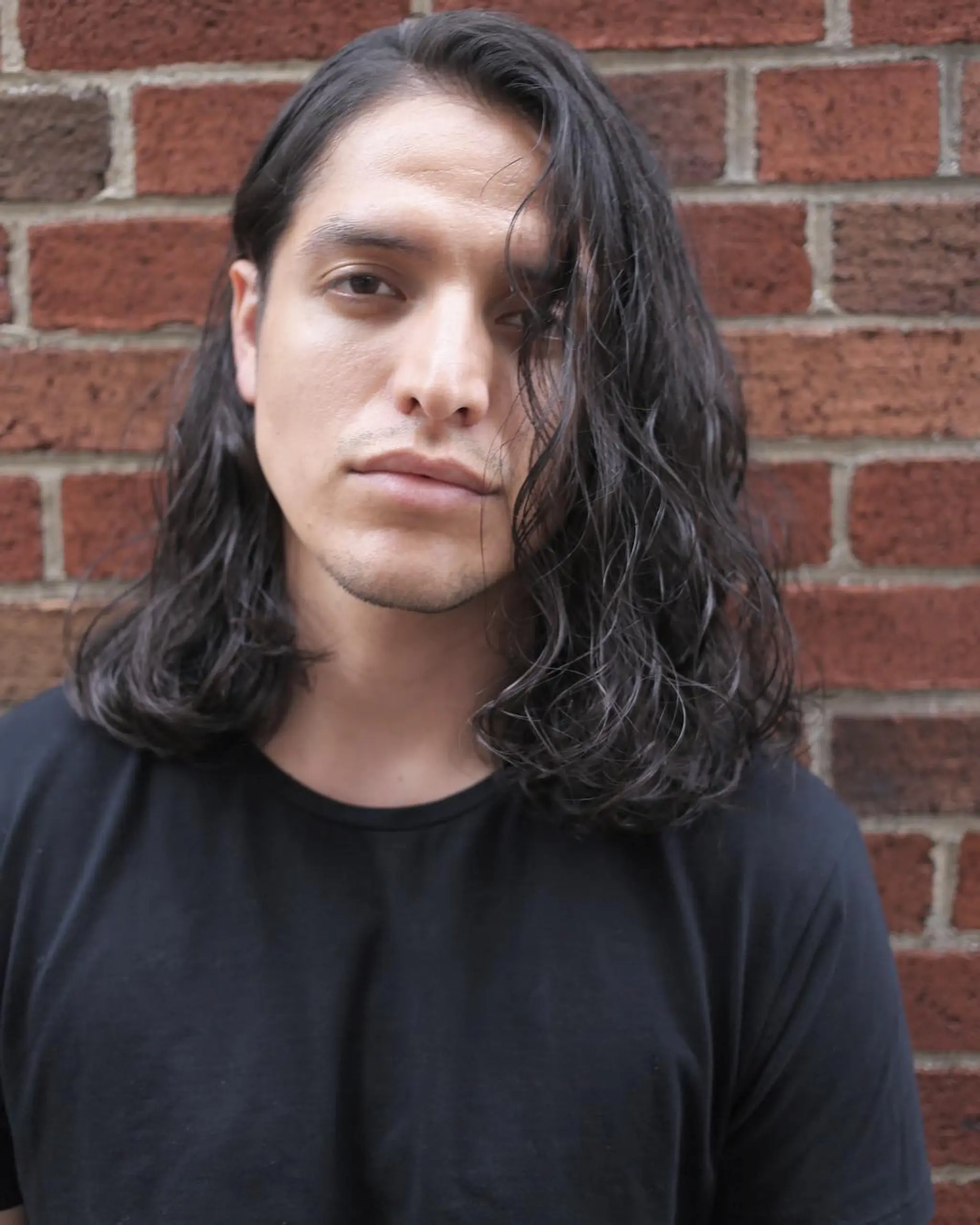 Medium-length wavy black hair with an asymmetrical flow, slightly off-center part, and neatly trimmed edges, creating a casual, laid-back vibe.