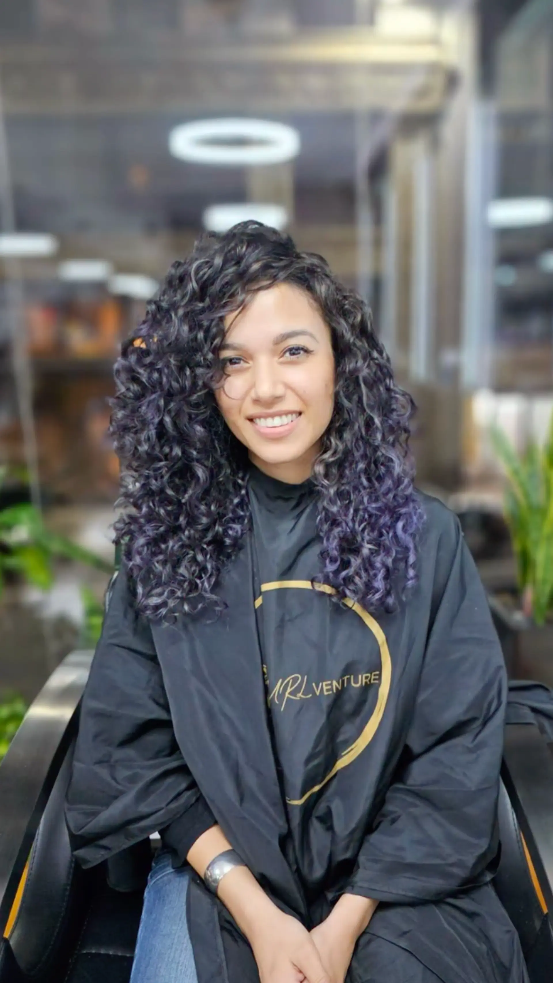 The hair features medium-length defined curls with a natural volume, transitioning from a black base color to vibrant purple highlights, forming a root-to-end gradient. 1