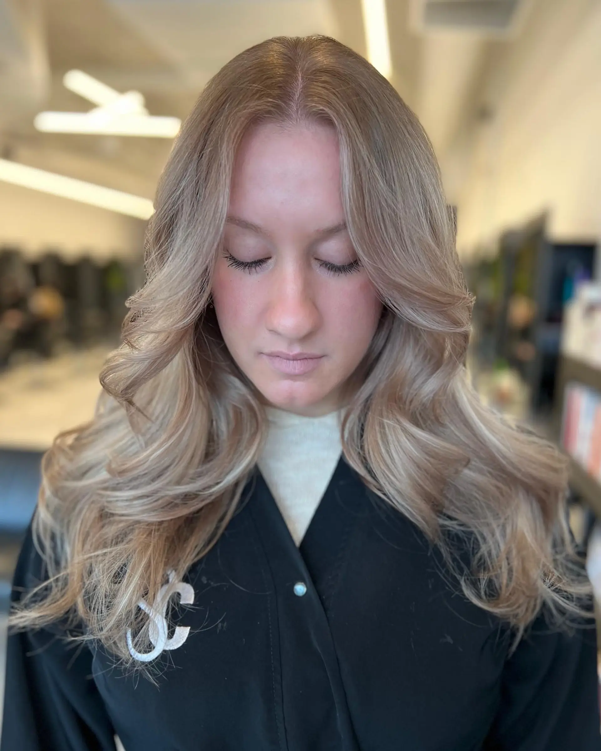 Long beige blonde hair styled with soft, voluminous waves and sweeping curtain bangs parted down the middle. 1