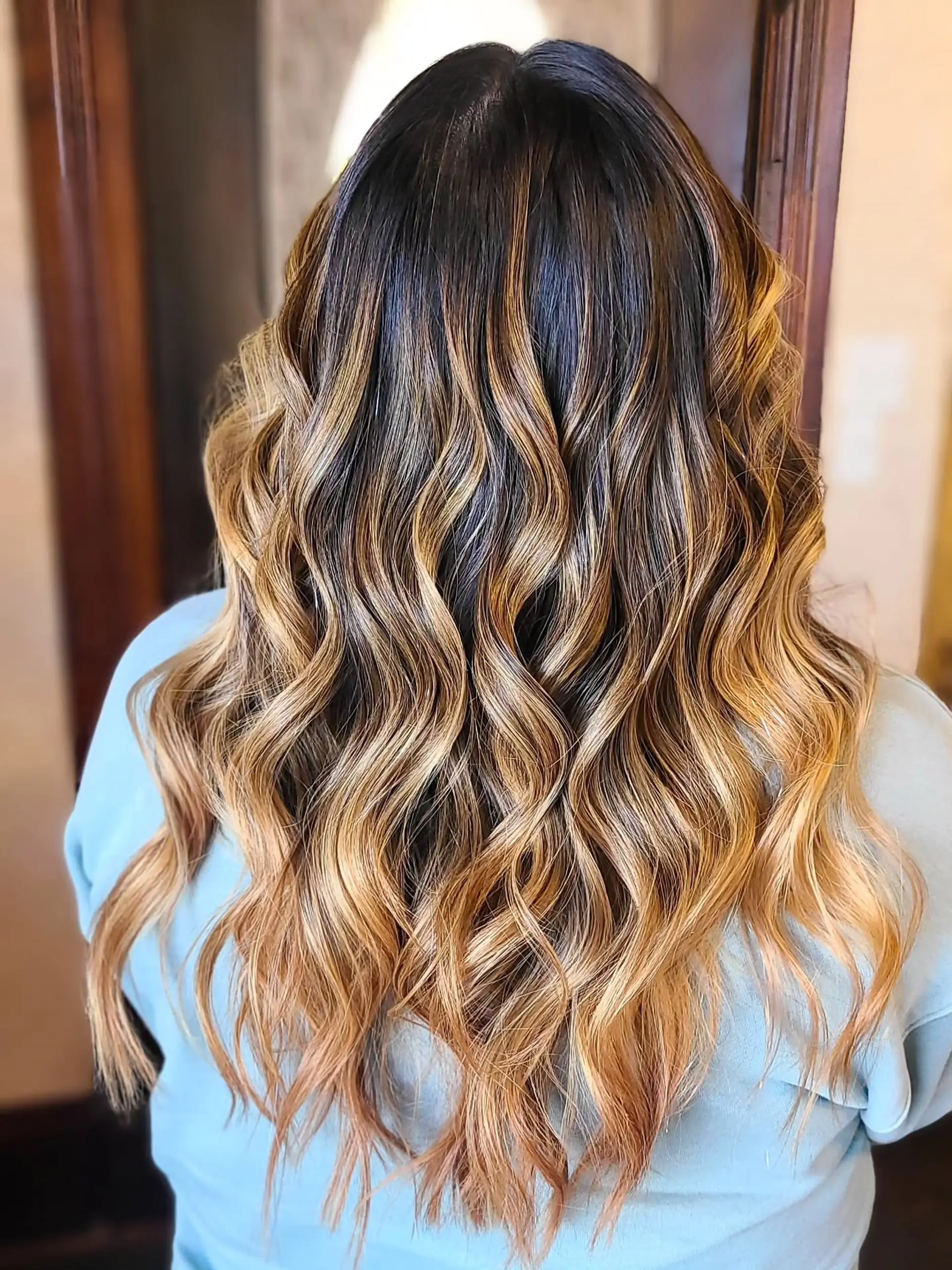 Long, wavy hair with a blended balayage coloring technique that transitions from dark roots to warm caramel and golden blonde ends, styled with soft, loose waves. 2