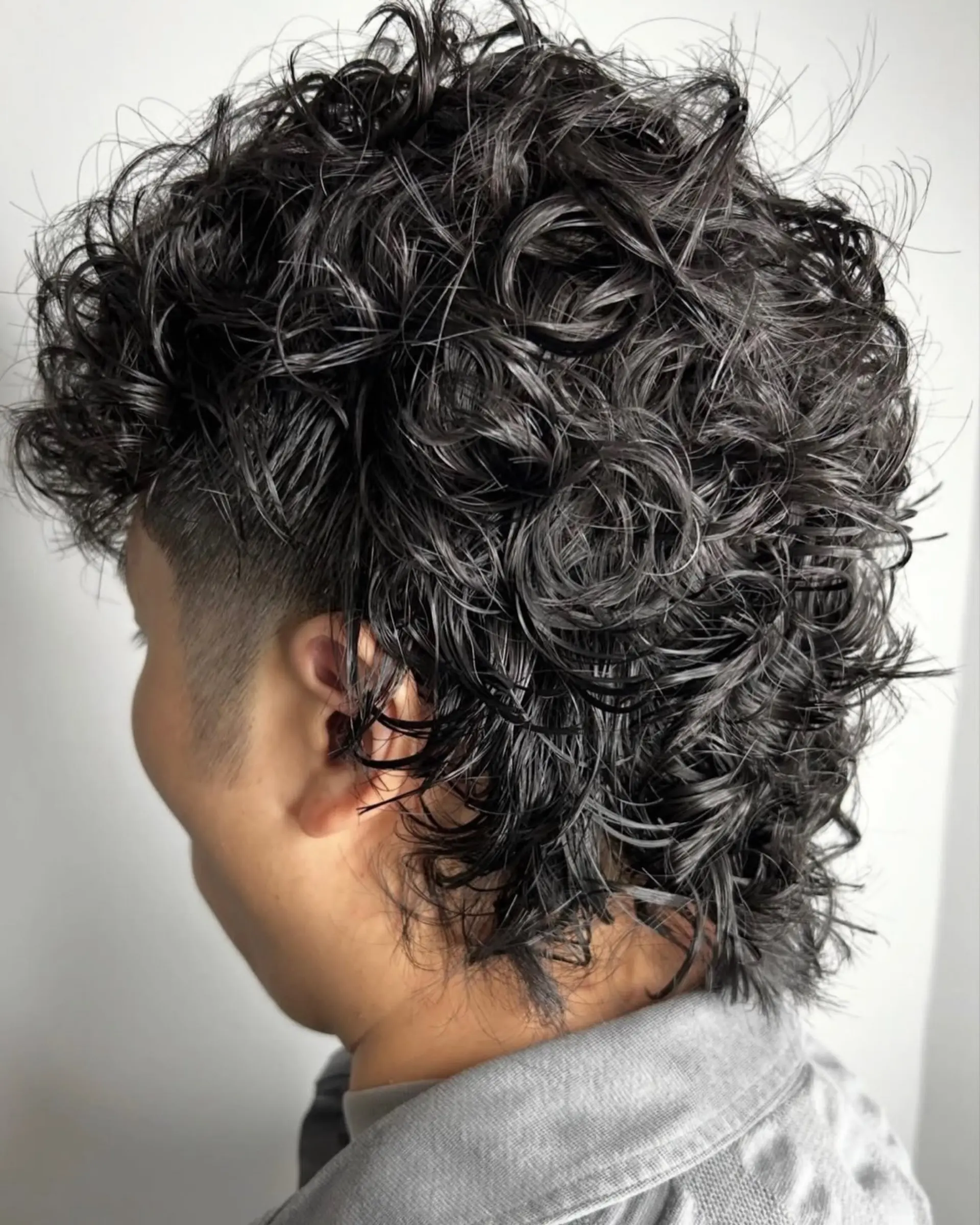 A modern curly mullet featuring tight permed curls throughout the top and back with tapered, faded sides around the ears. 1