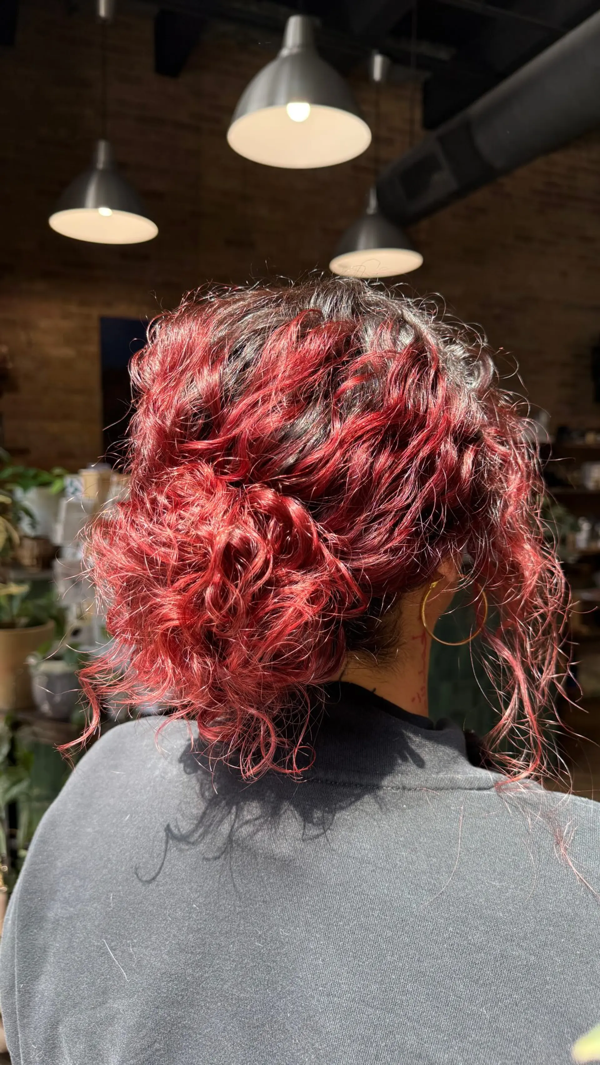 A textured, messy low bun featuring curly hair with vibrant crimson-red color over dark roots and loose face-framing tendrils. 1
