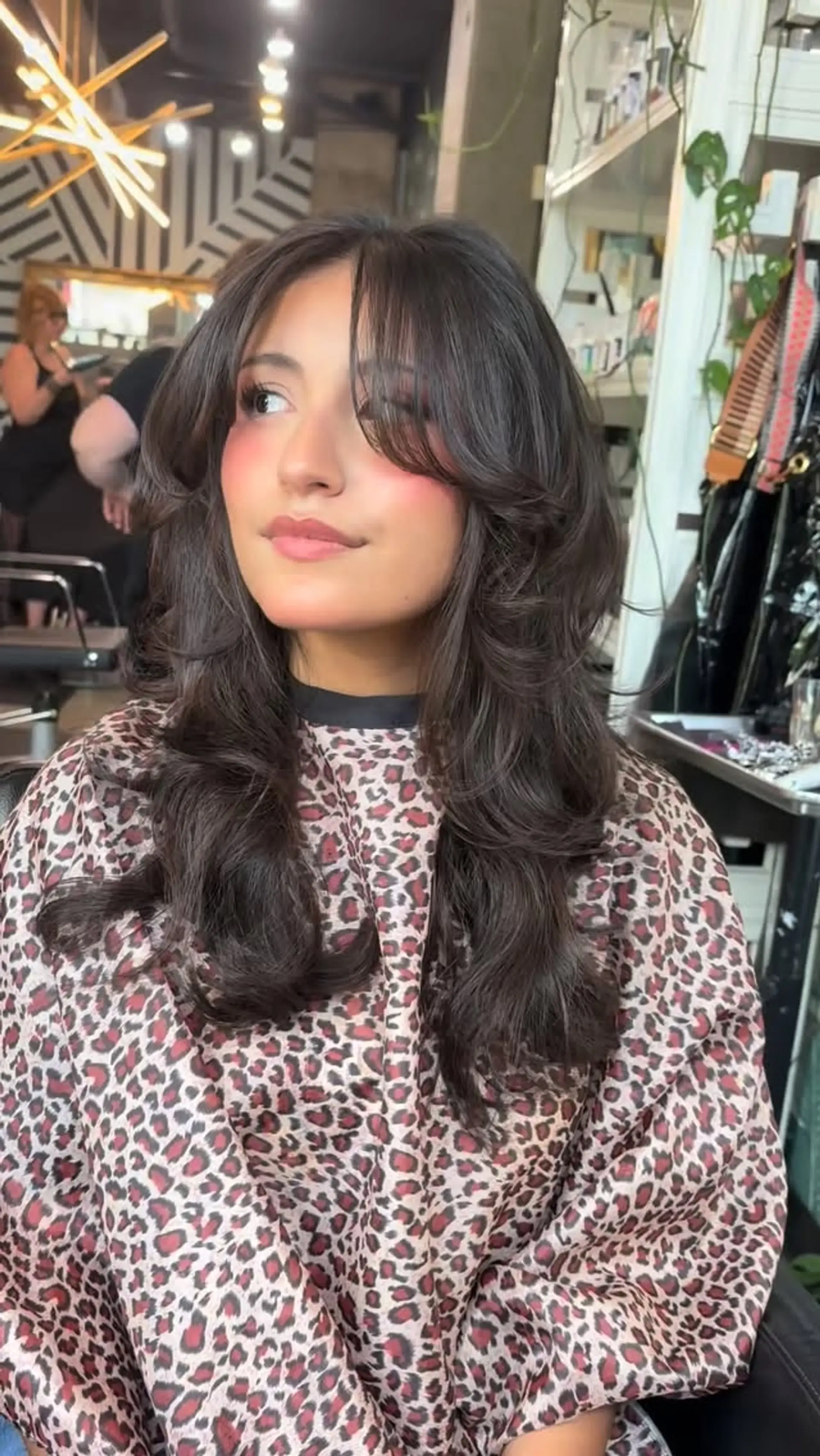 Long dark brown hair cut with abundant face-framing layers, wispy curtain bangs, and bouncy outward curls. 1