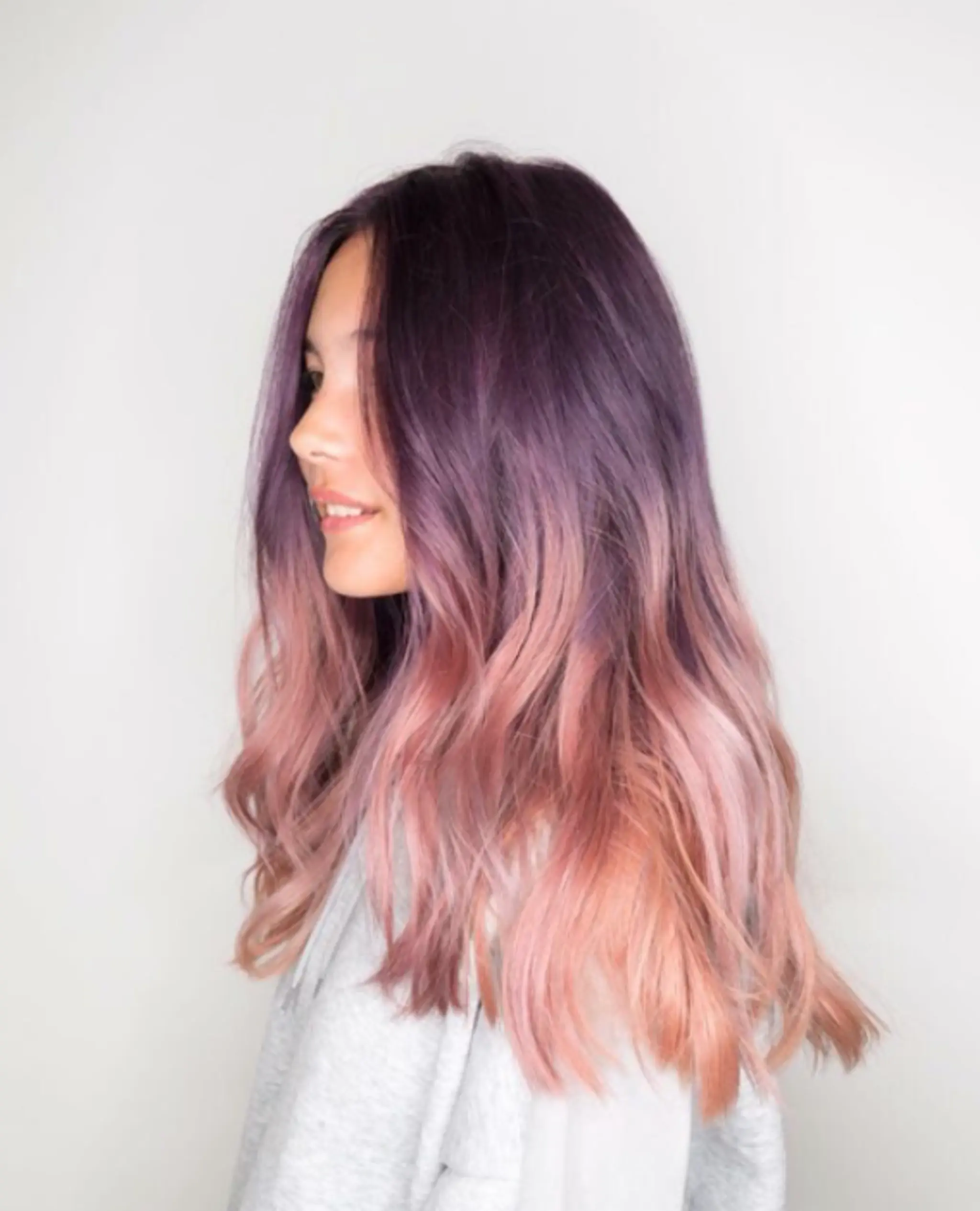 A long wavy hairstyle with an ombre effect transitioning from deep purple at the roots to lavender mid-section, and ending with soft pink and blonde tips. The waves are soft and natural, complemented by medium layers for added texture.