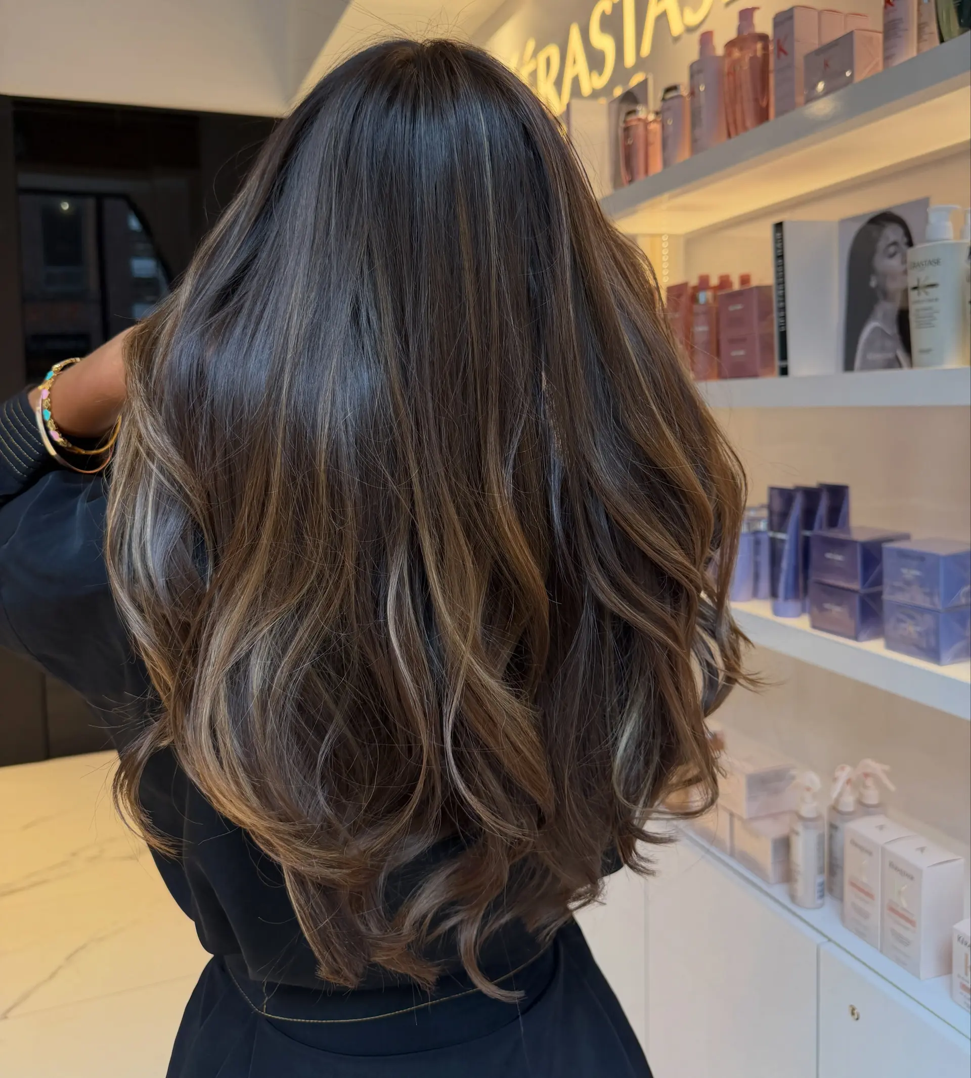 Long dark brunette hair enhanced with soft dimensional balayage highlights and styled into voluminous waves. 1