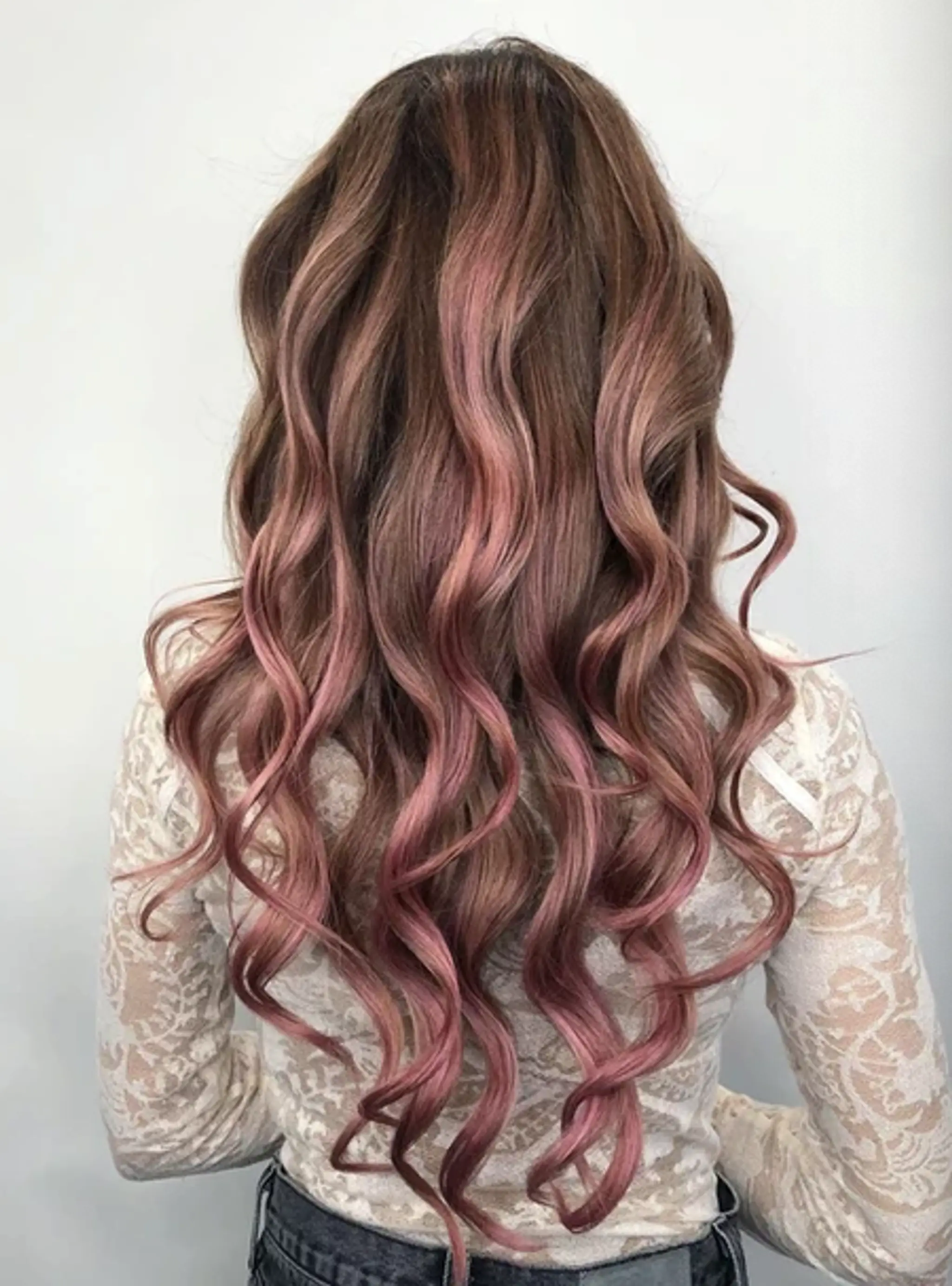 Long, wavy hair with a foliage blend of brown roots to rose gold ends, featuring loose waves and soft curls in layered style.