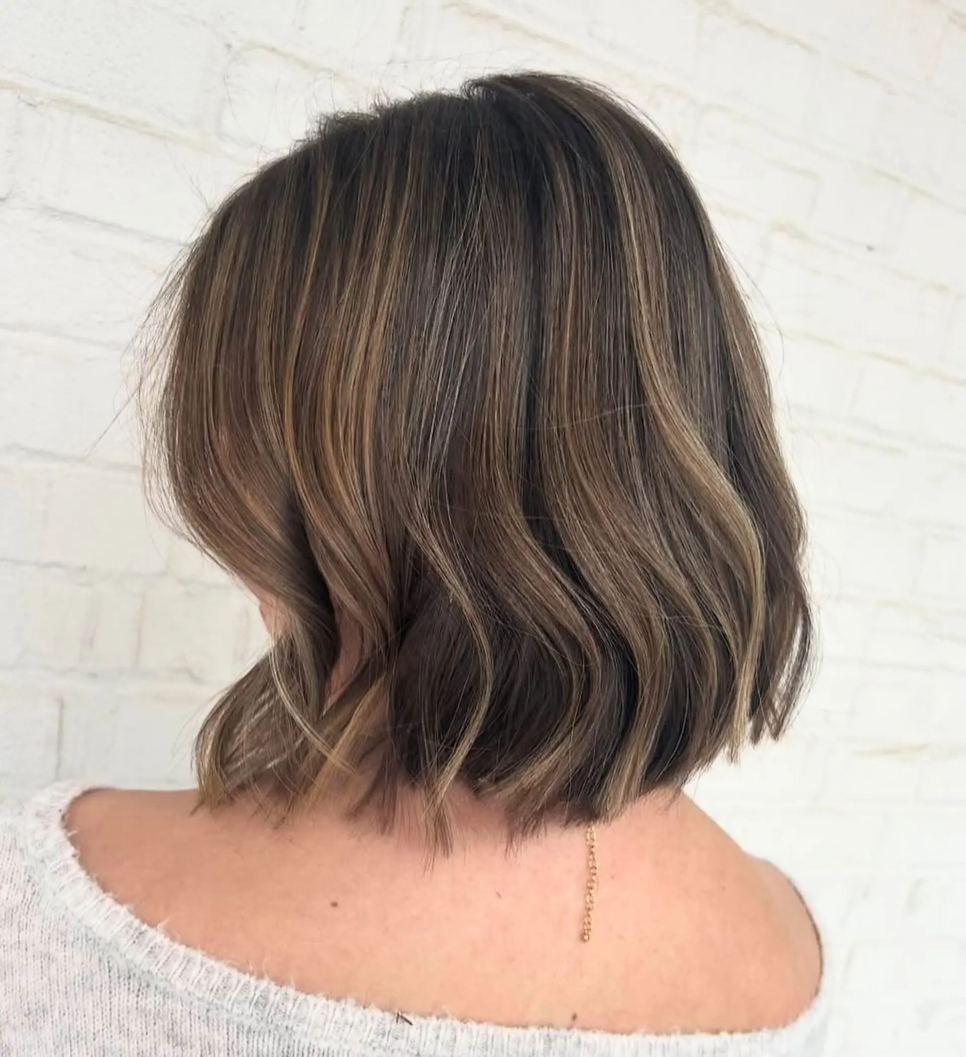 A blunt, textured chin-to-neck length bob styled with gentle bends and waves, featuring soft dimensional caramel highlights over a rich brunette base. 1