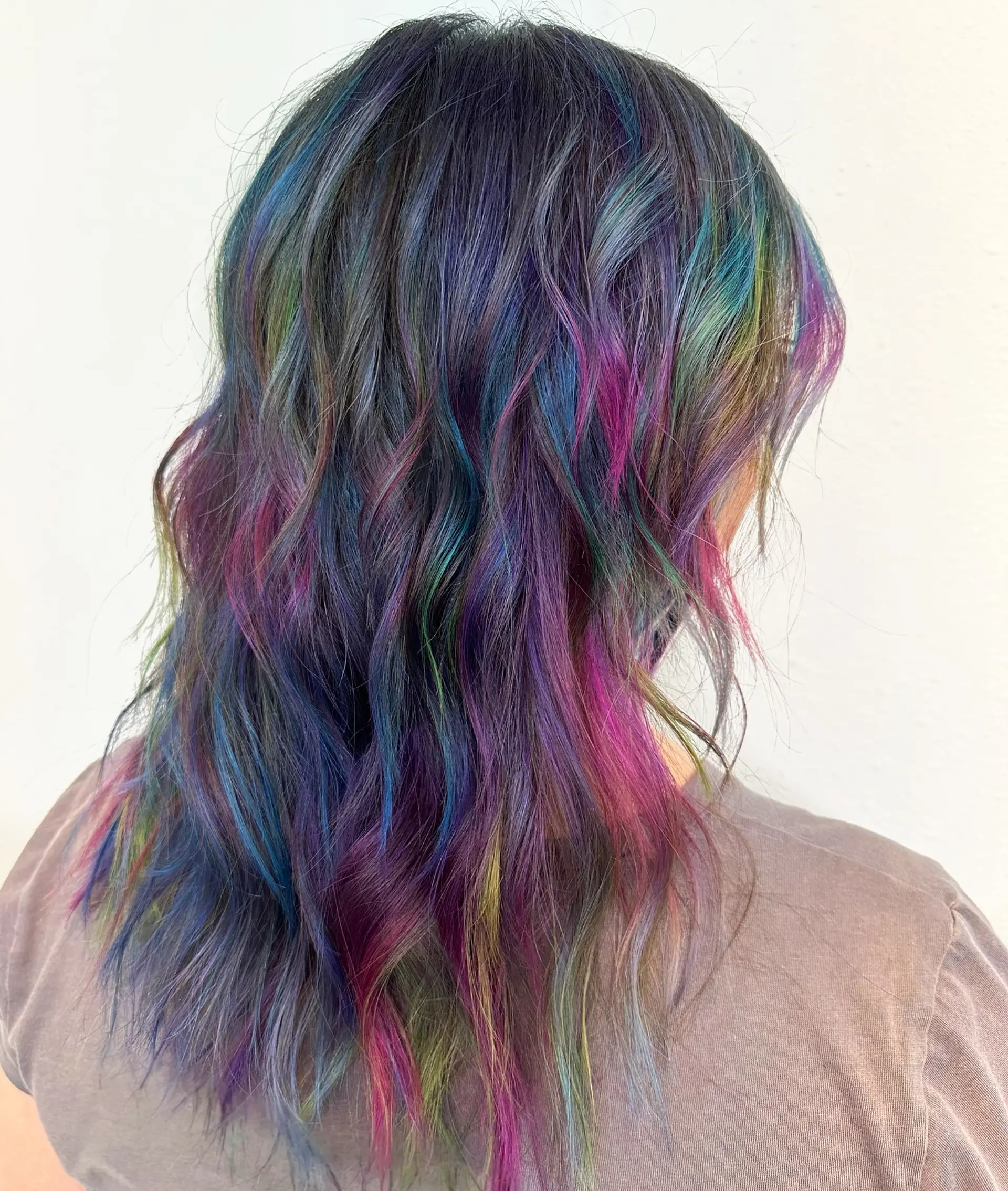 Medium-to-long wavy hair featuring an oil slick or galaxy color melt with tones of deep purple, blue, magenta, and green woven through a dark base. 1