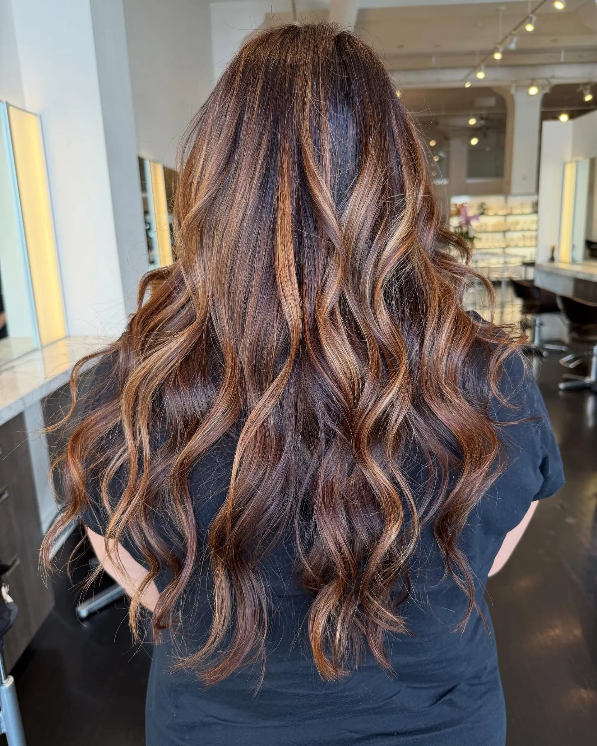 Long dark brunette hair featuring warm caramel highlights and styled with loose, dimensional waves. 1