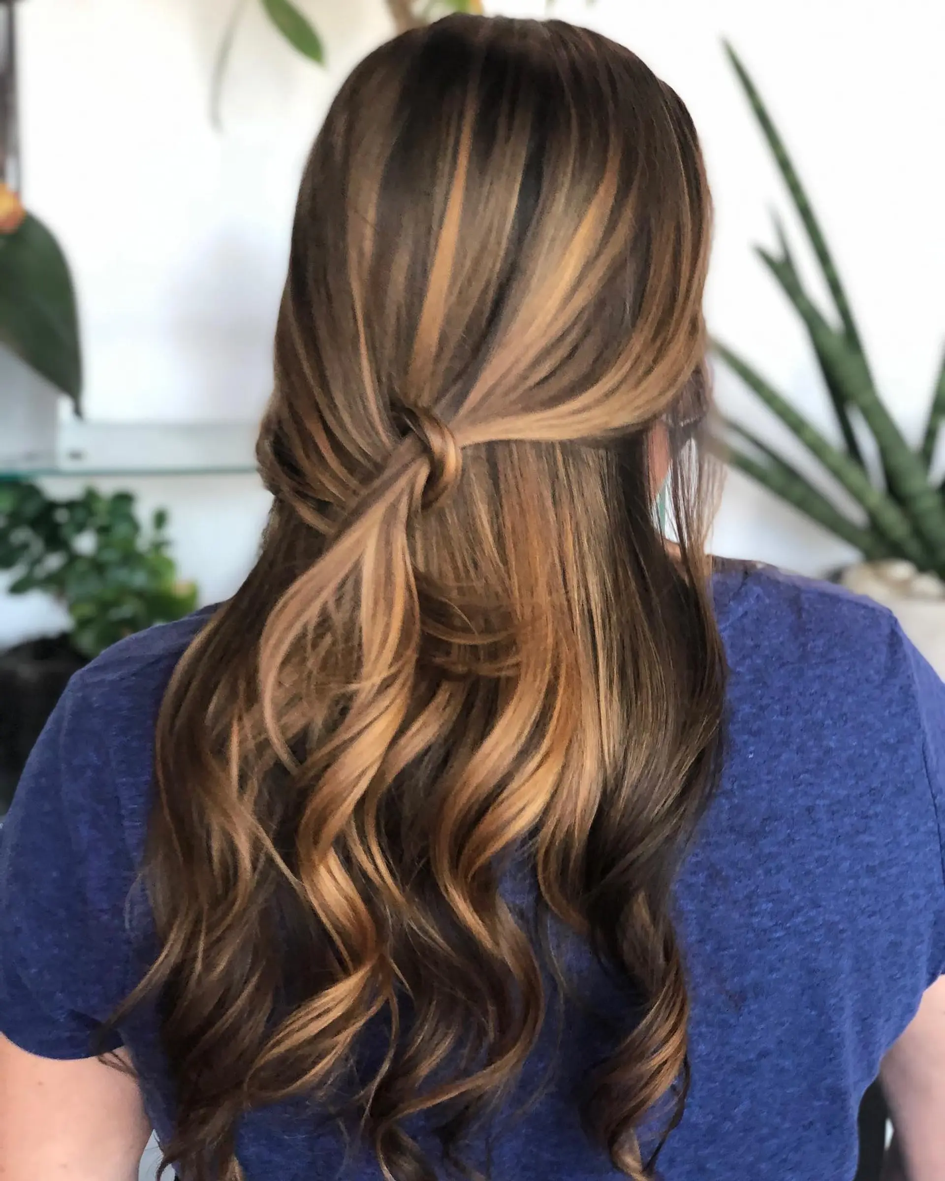 Long brunette hair with warm caramel balayage, gathered into a knotted half-up style with loose cascading waves. 1