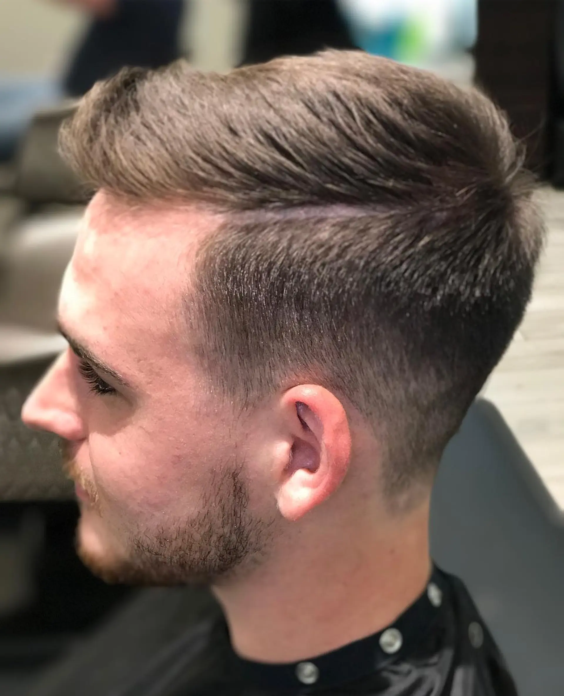 A men's short taper fade haircut with a defined side part and textured volume swept back on top. 1