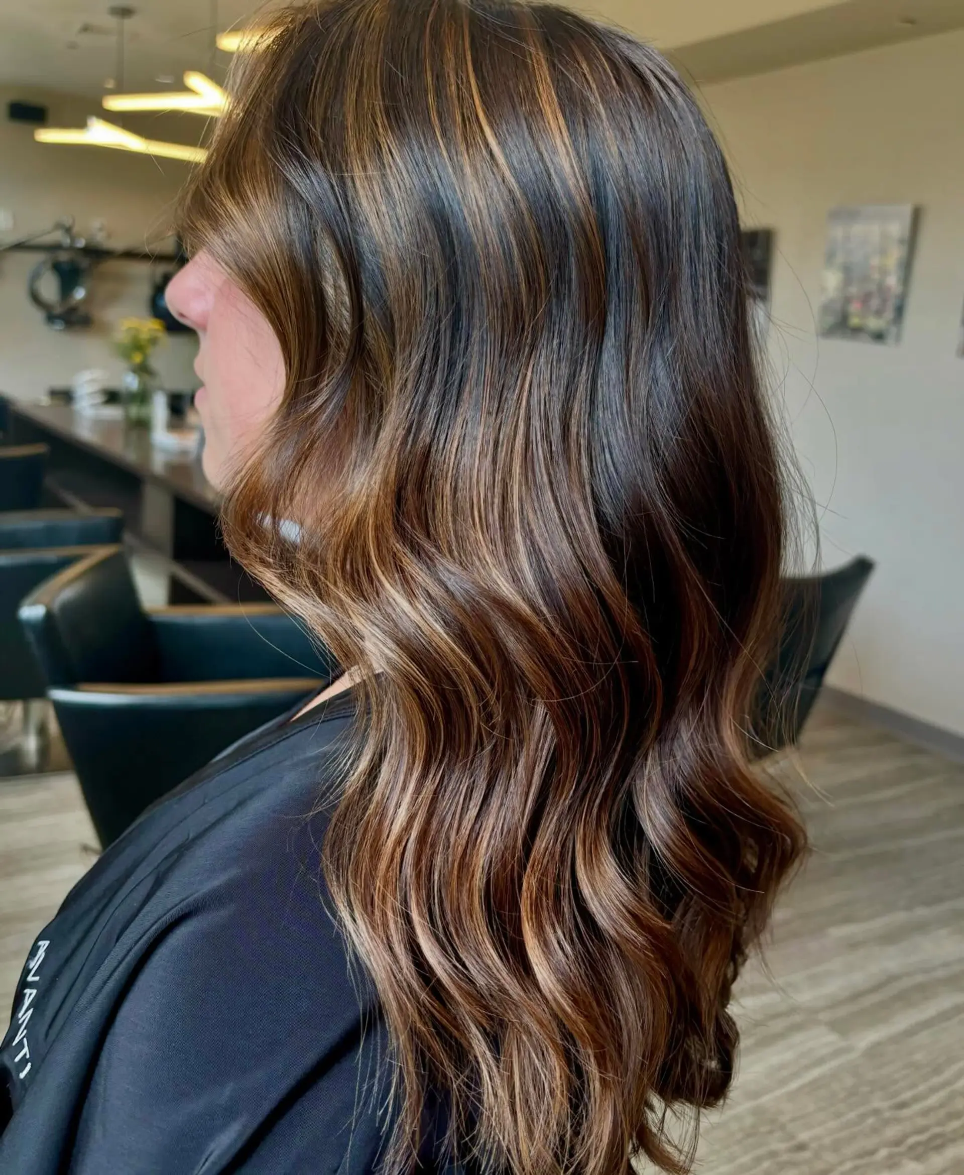 Long dark brunette hair featuring dimensional warm caramel balayage highlights styled in glossy, soft waves. 1