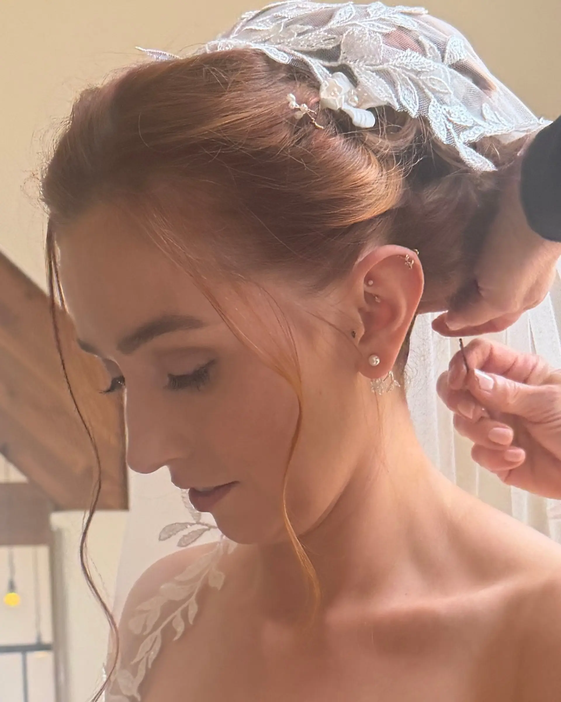An elegant bridal updo adorned with a delicate lace veil, decorative hairpin, and loose face-framing tendrils. 1