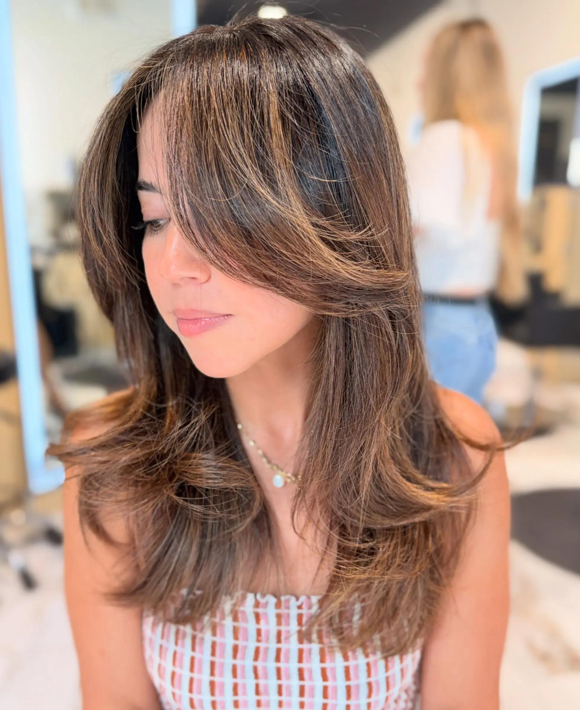Long layered brunette hair featuring subtle caramel highlights and a voluminous blowout with soft, face-framing curtain bangs. 1