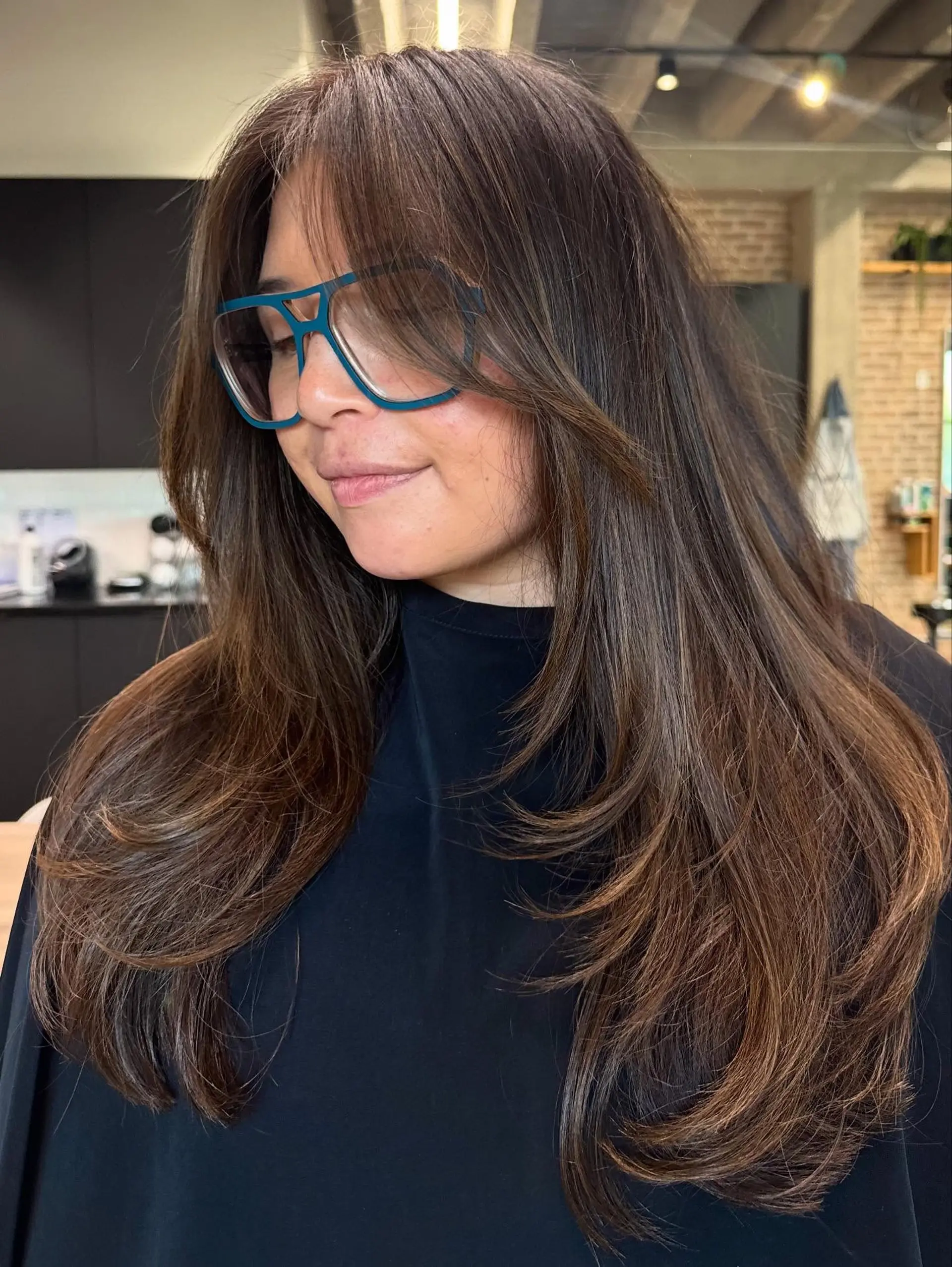 This long layered cut features extensive face-framing layers and curtain bangs, creating soft volume and movement throughout the hair. The ends are texturized and appear bouncy. 2