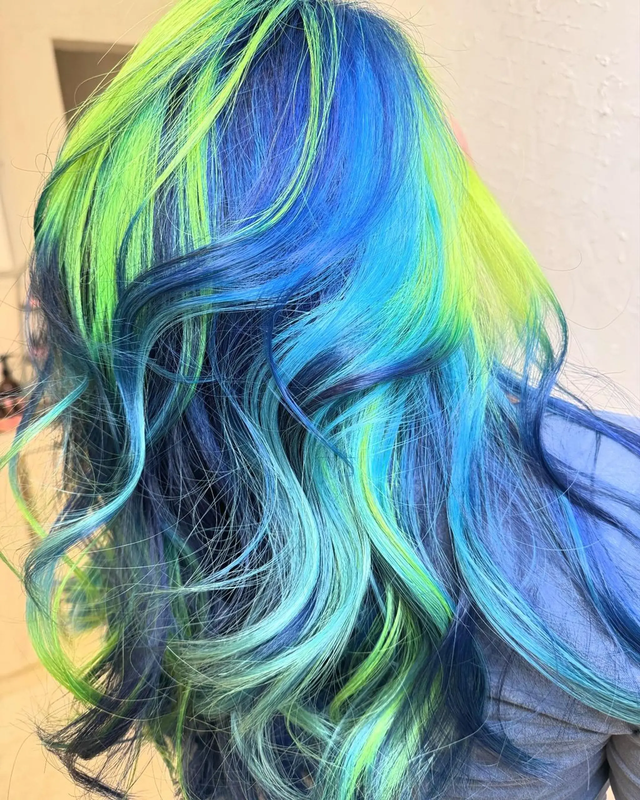 Long, wavy hair with a vibrant blend of neon green and electric blue hues, styled in loose waves for a voluminous, playful appearance.