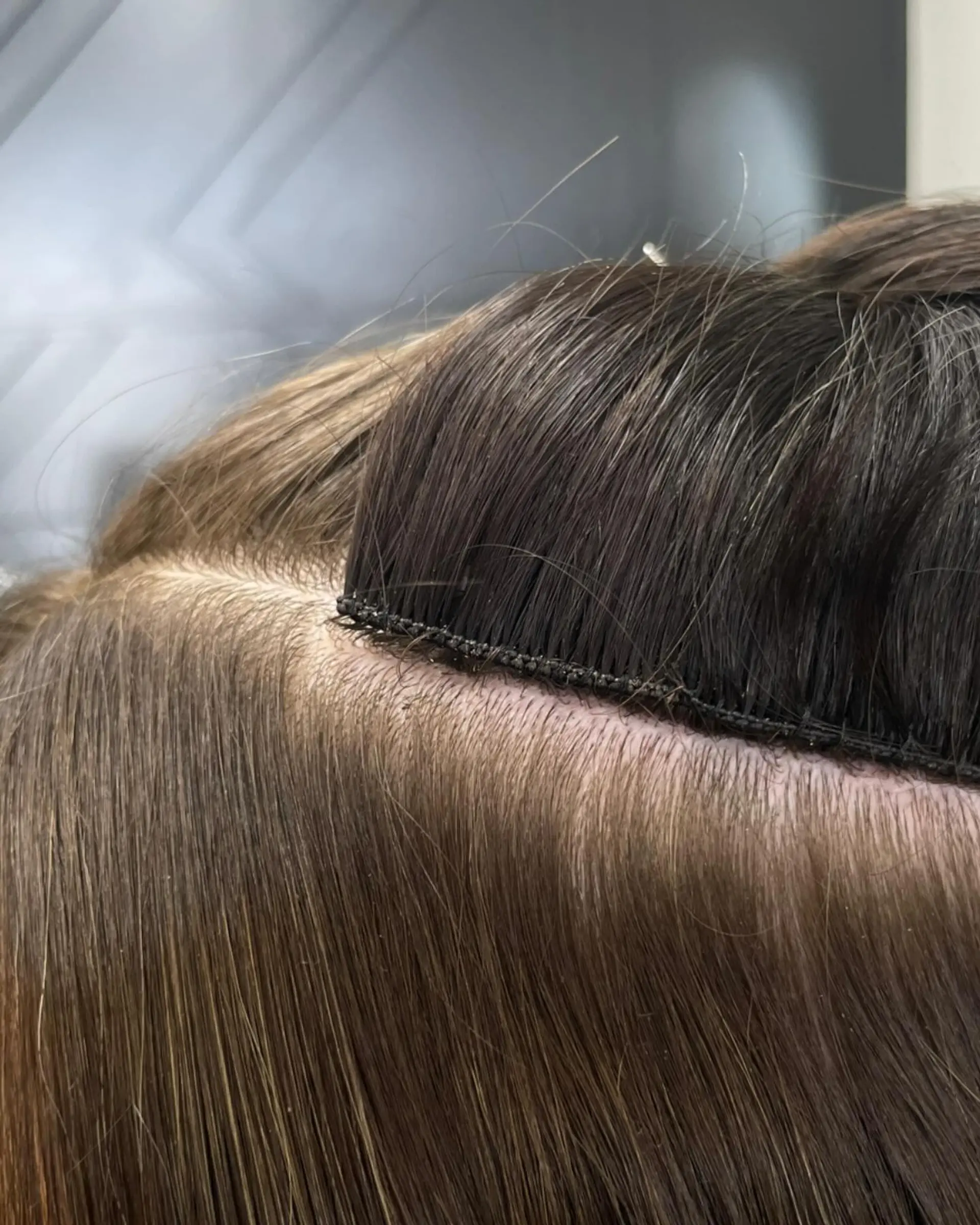 A close-up view displaying the neat and seamless seam of a hand-tied hair extension row applied to brunette hair. 1