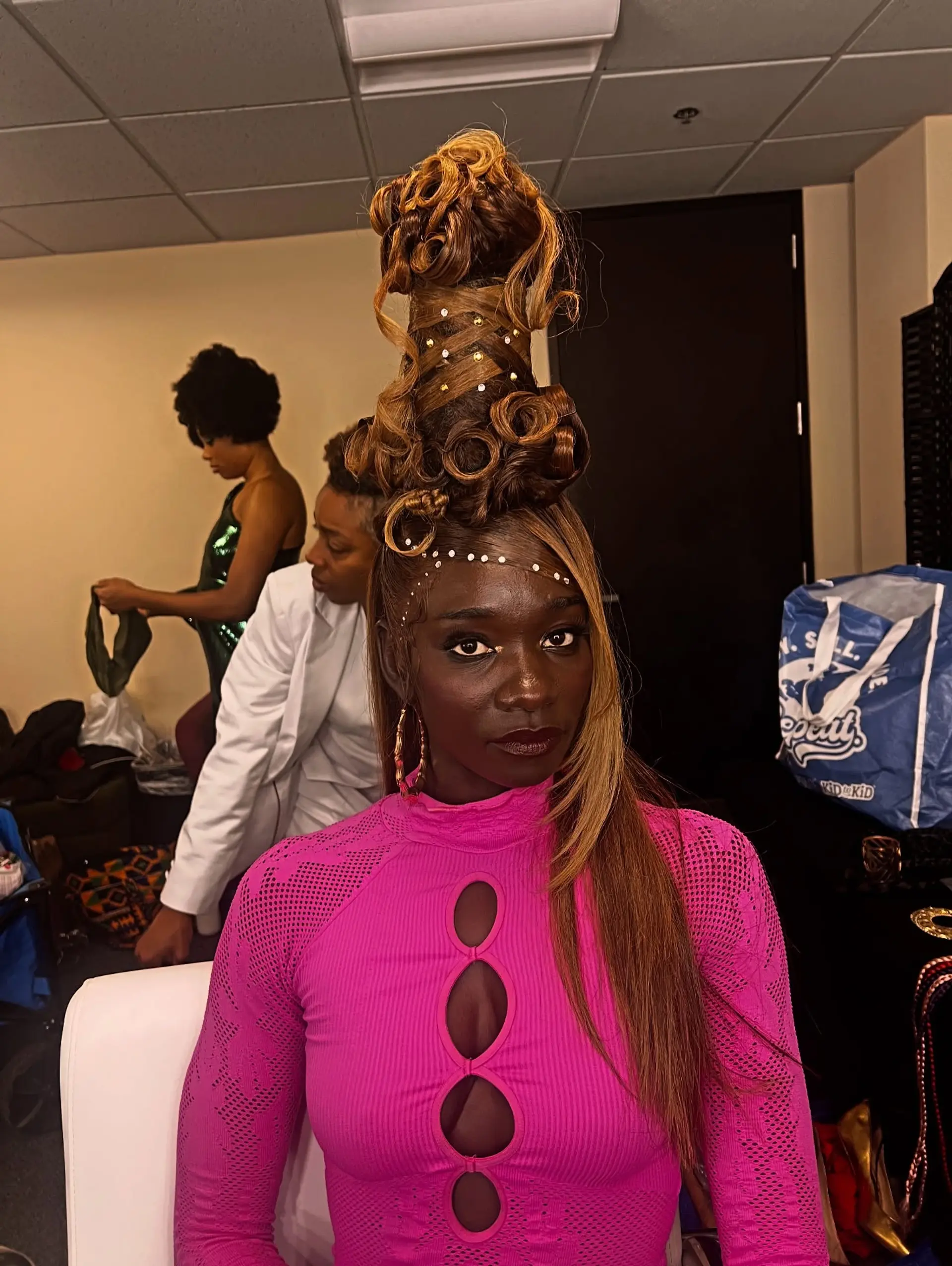 A dramatic, vertical sculpted tower updo featuring intricate loops, curls, and rhinestone embellishments with a sleek side piece. 1