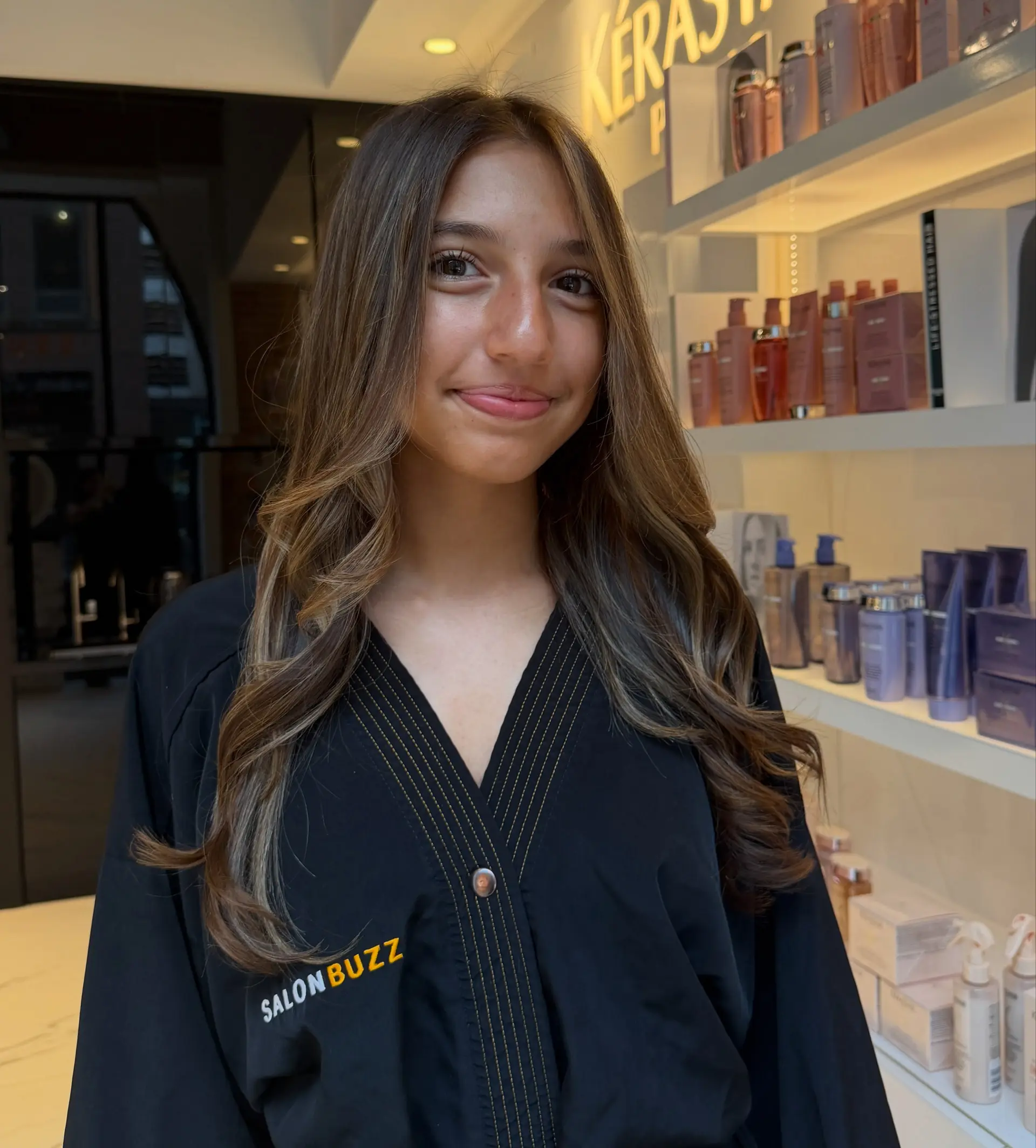 Long brown hair enhanced with soft caramel highlights and styled into loose, face-framing waves. 1