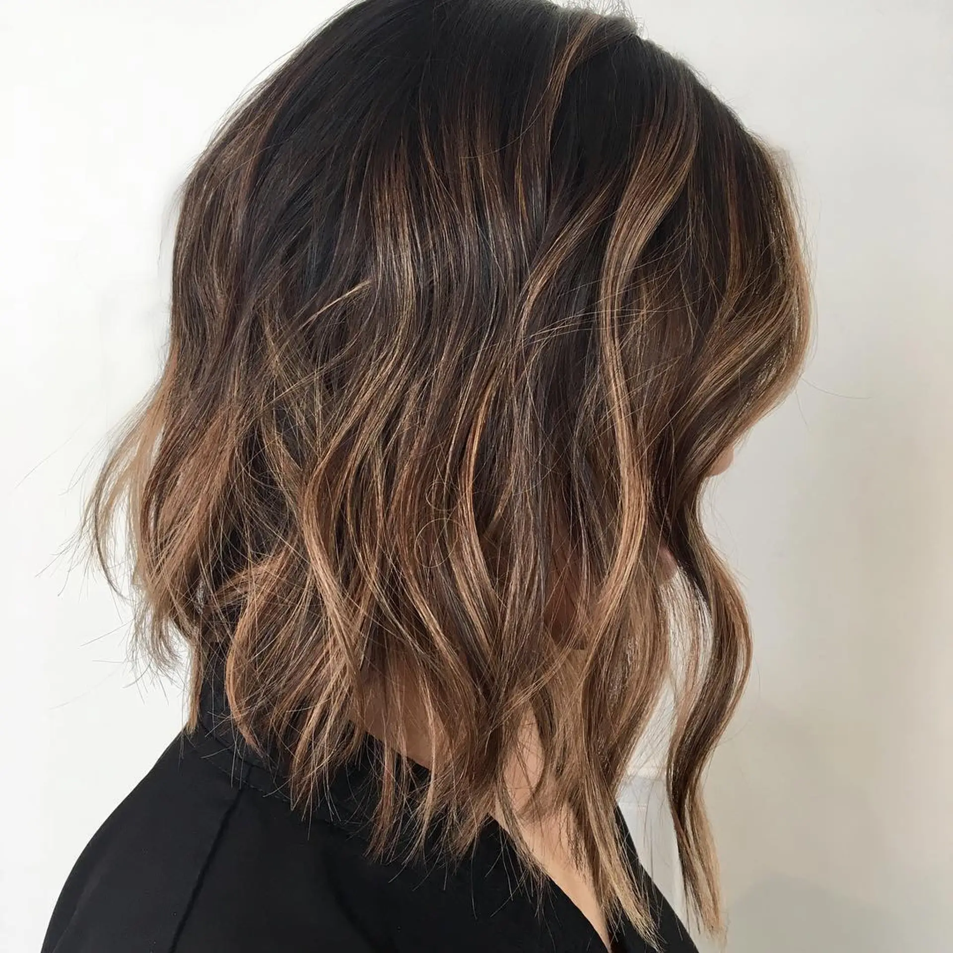 A textured angled lob styled in loose waves, highlighted with dimensional caramel and warm brown tones over a dark base. 1