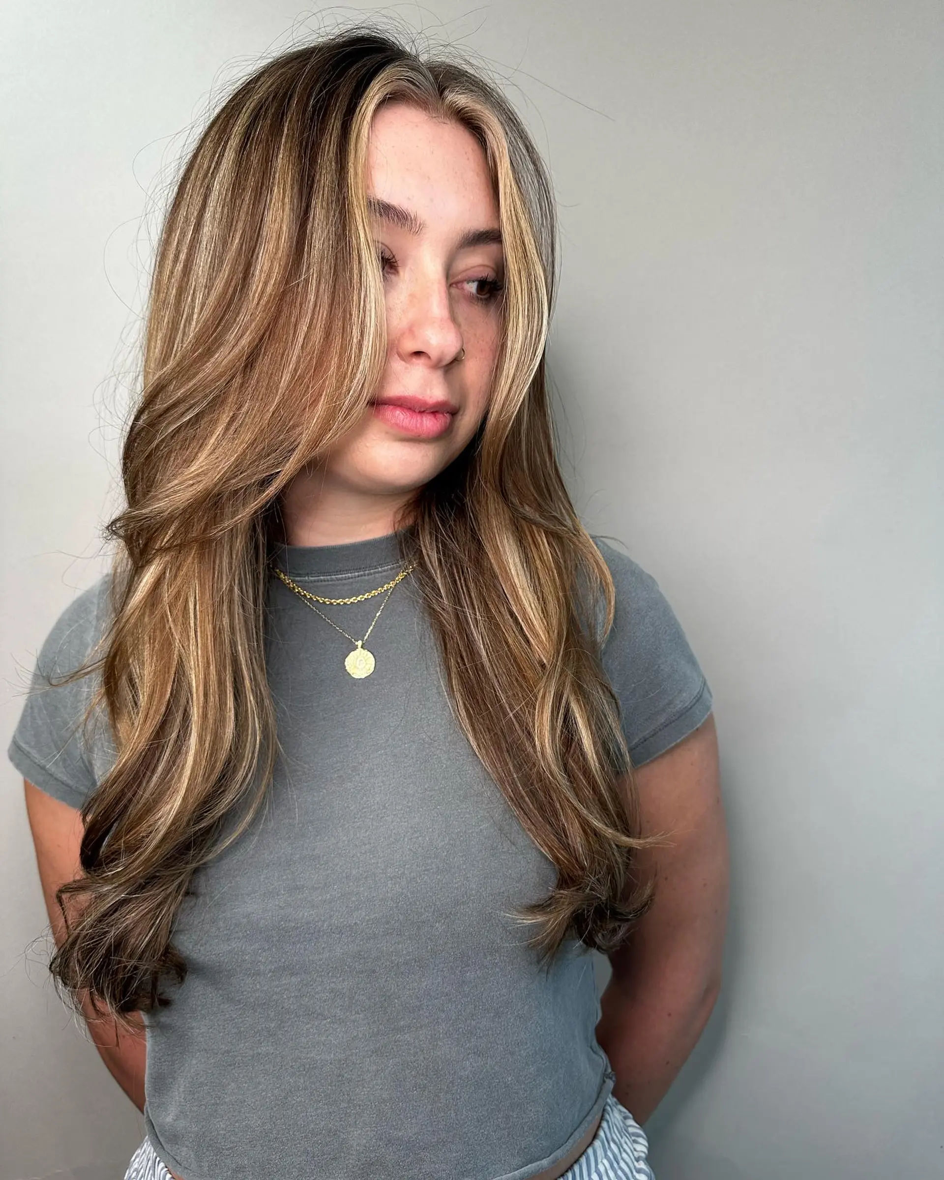 Long dimensional brunette hair with soft golden blonde highlights, bright face-framing money pieces, and gentle blowout waves. 1
