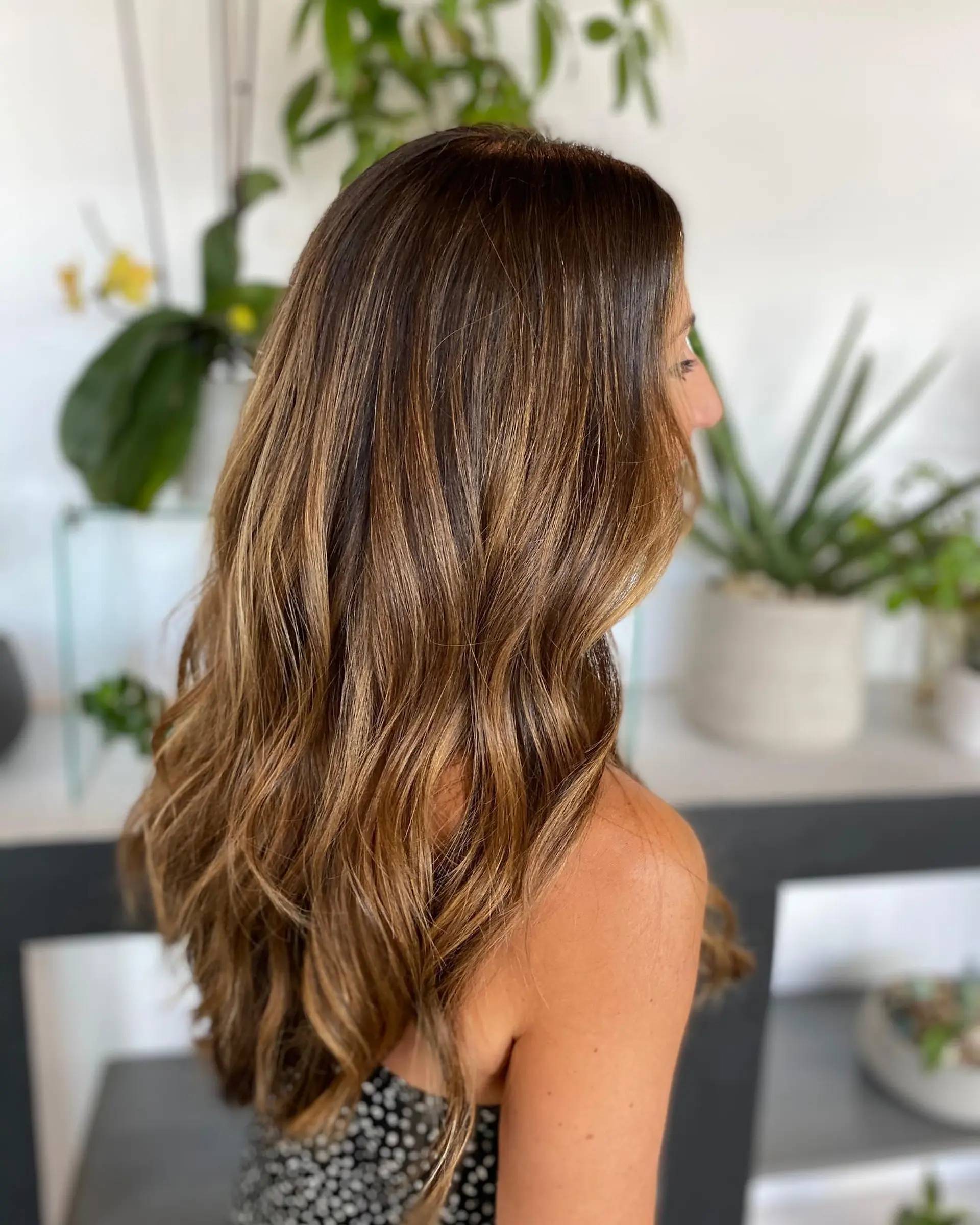 Long dimensional brunette hair styled in loose waves, featuring blended caramel and golden balayage highlights alongside rich lowlights. 1