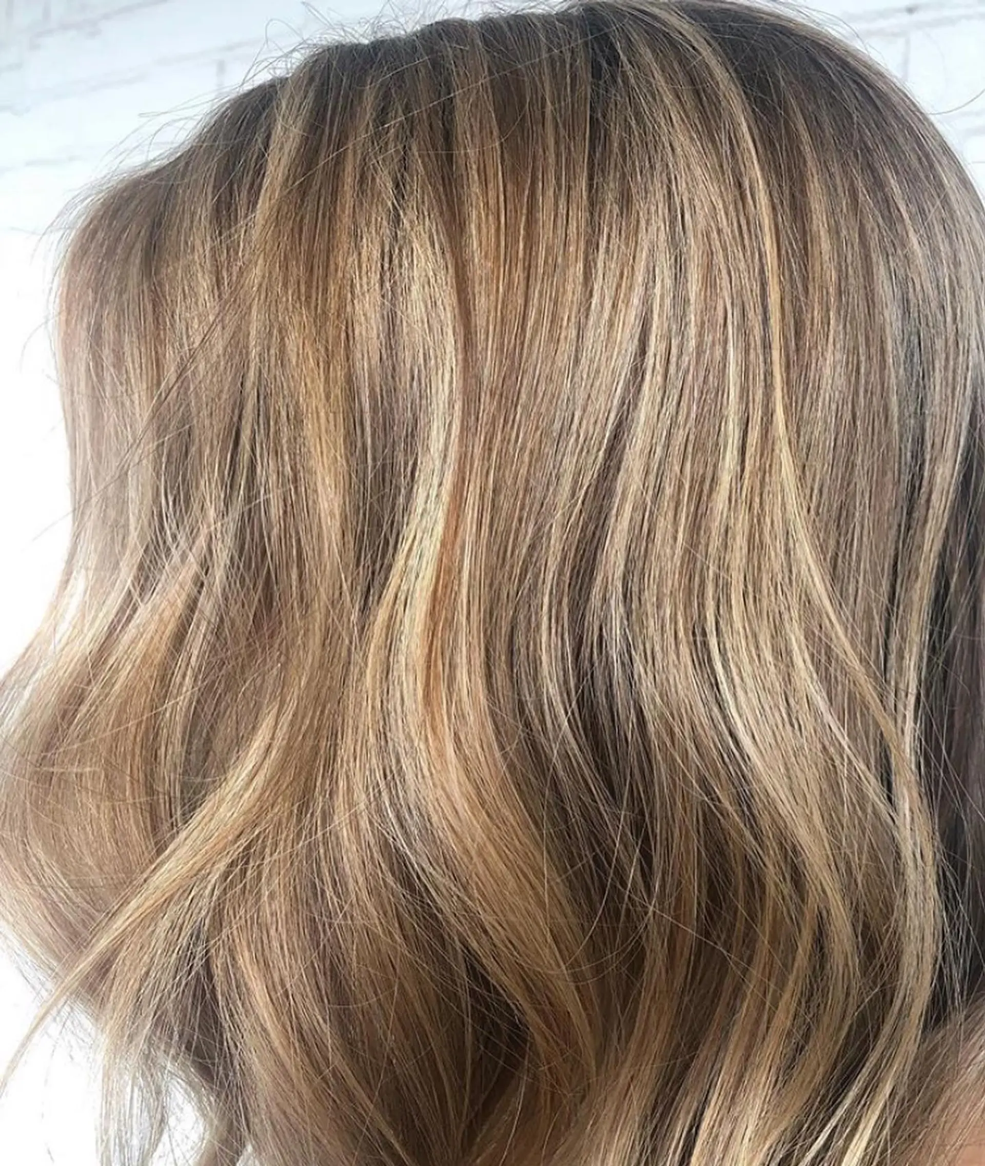 Soft dimensional warm blonde highlights blended seamlessly through light brown hair styled with loose, gentle waves. 1