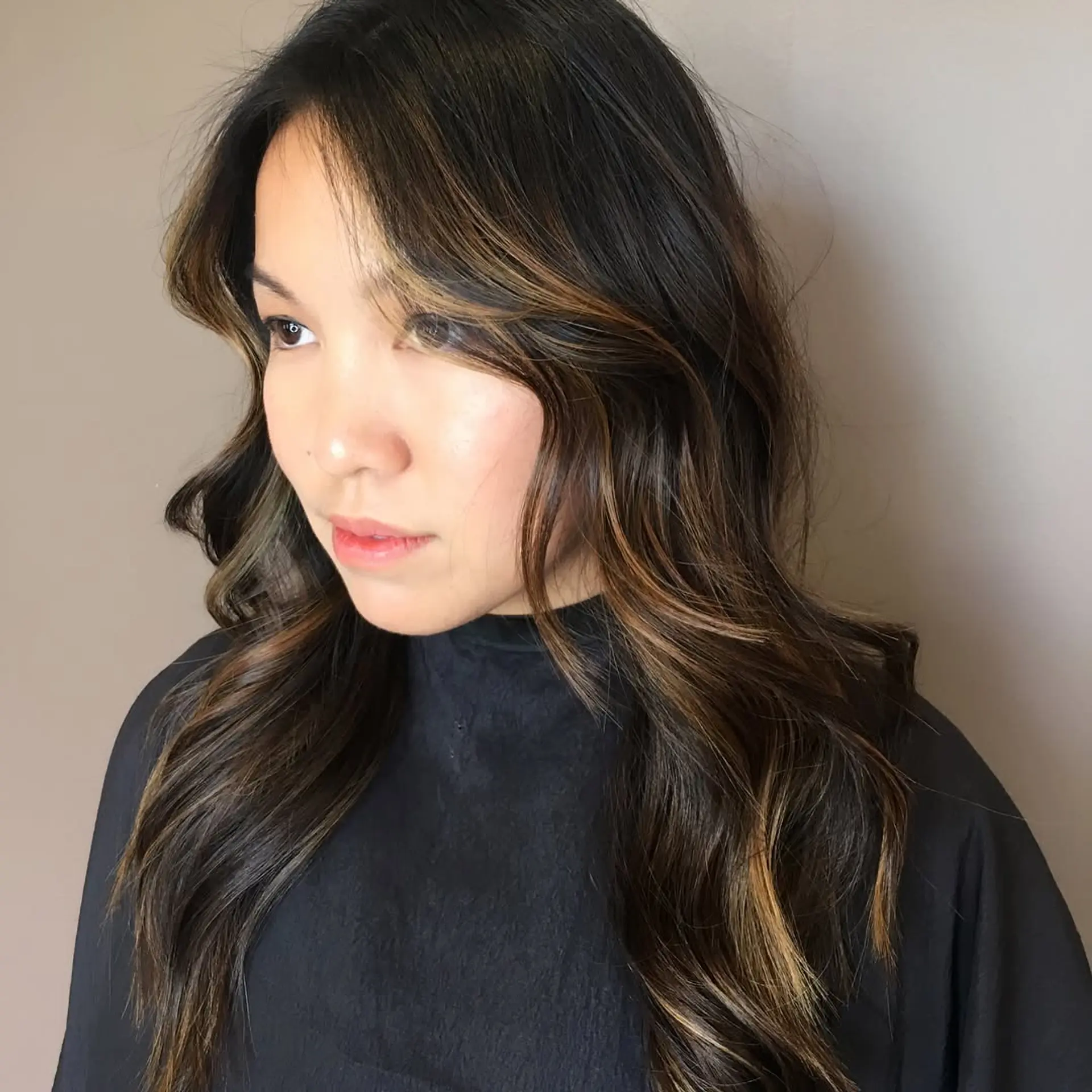 Long dark brown hair styled in soft, loose waves with subtle warm caramel balayage highlights framing the face. 1