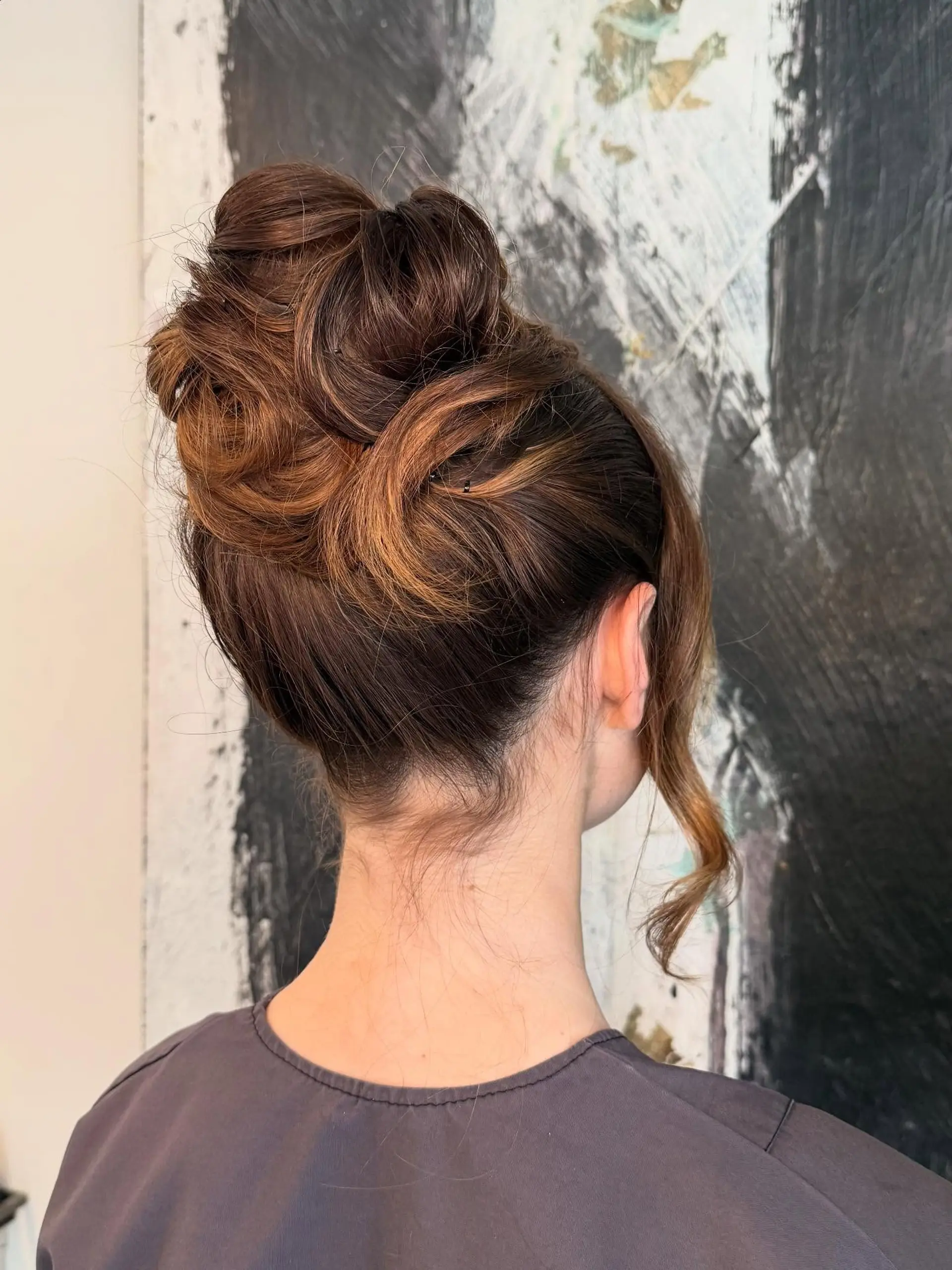 A sophisticated high updo styled with sculpted loops and voluminous curls, featuring warm caramel dimensional highlights and a soft face-framing tendril. 1