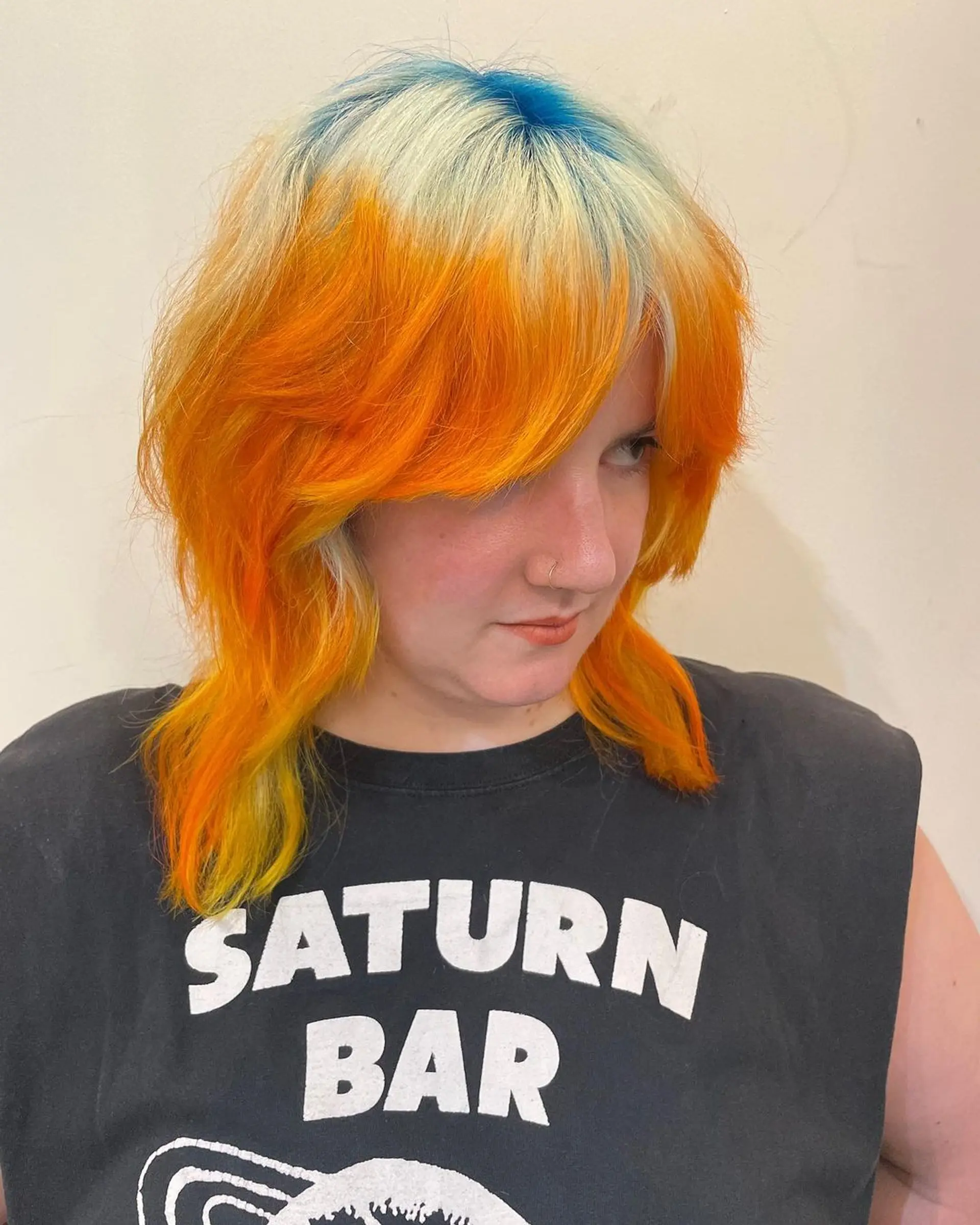 A vibrant and edgy medium-length wavy cut featuring bold orange, blonde, and blue hues. It includes textured waves and a face-framing fringe, with a distinct layered cut for added dimension. 1