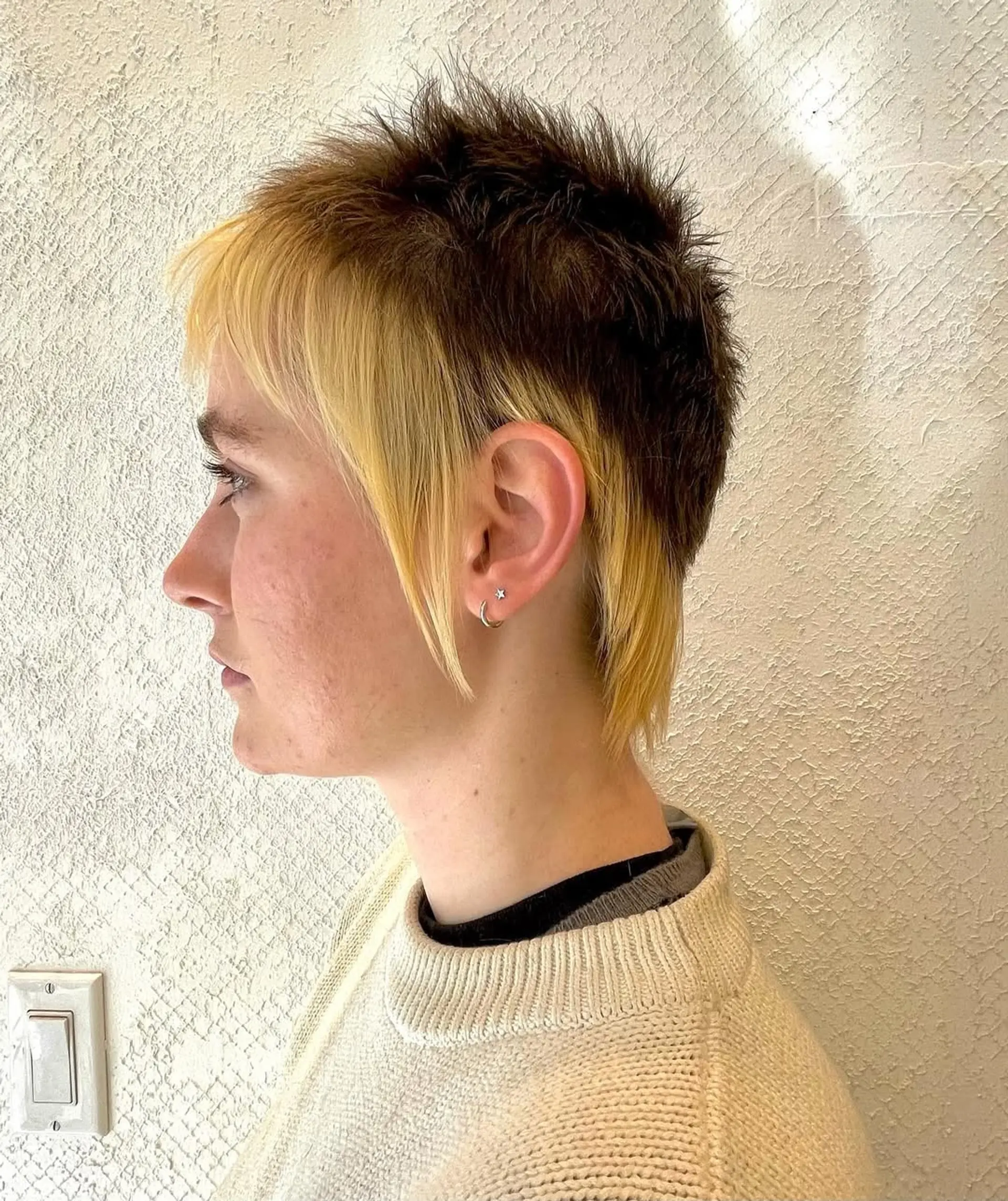A short, choppy mullet with a blonde and dark brown contrast. The hairstyle features an undercut, a textured top, and an ear-length fringe with straight, layered strands for a spiky look. 1
