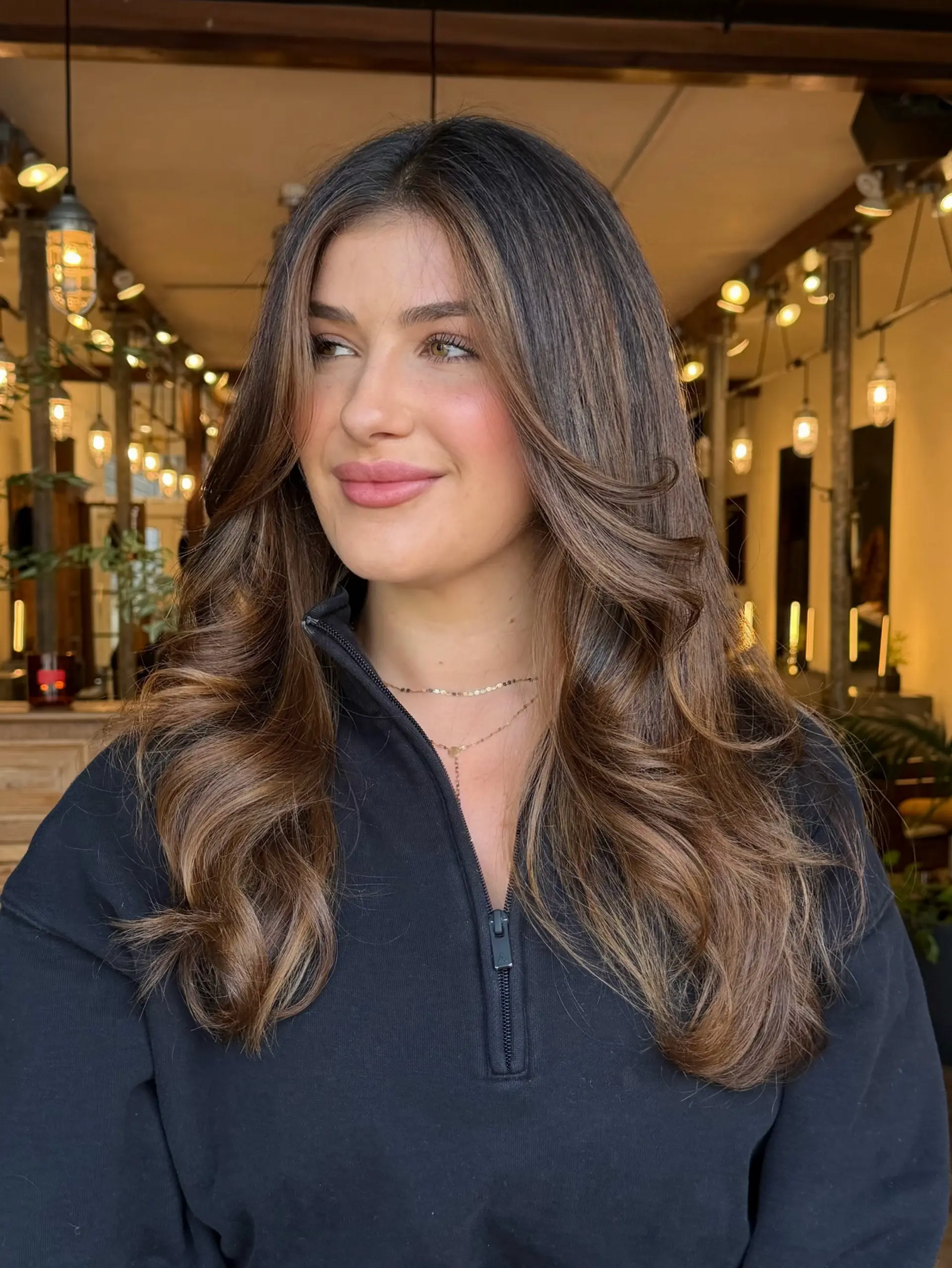 Long layered warm brunette hair styled with voluminous, bouncy waves and sun-kissed dimensional balayage highlights. 1