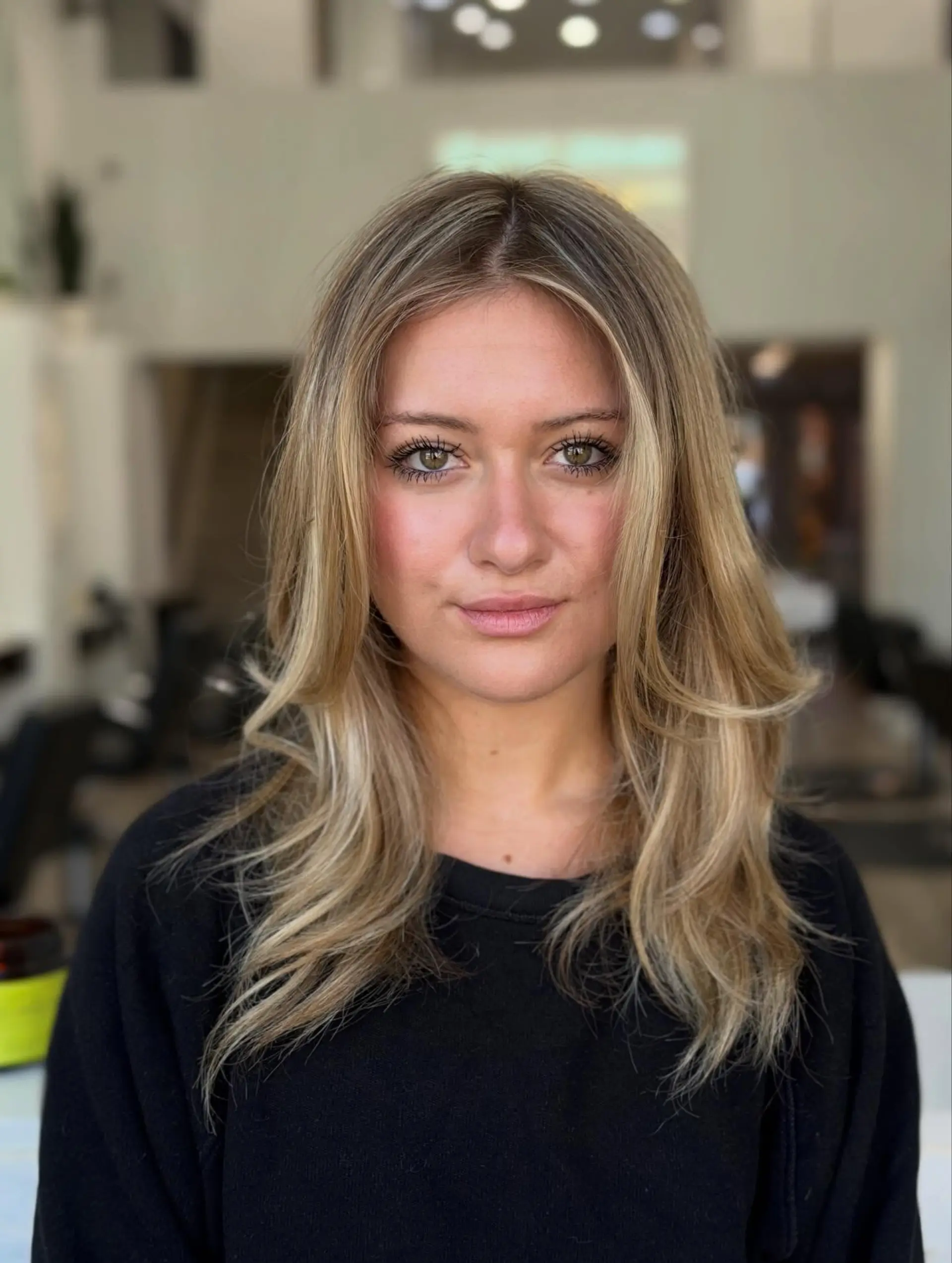 Medium-length blonde hair styled with soft, face-framing curtain layers and feathered ends. Estimate scope: Haircut and styling only. Blonde highlights in the description are not included in this mapped estimate. Published salon-level starting/range estimate; confirm the chosen provider’s quote. 1