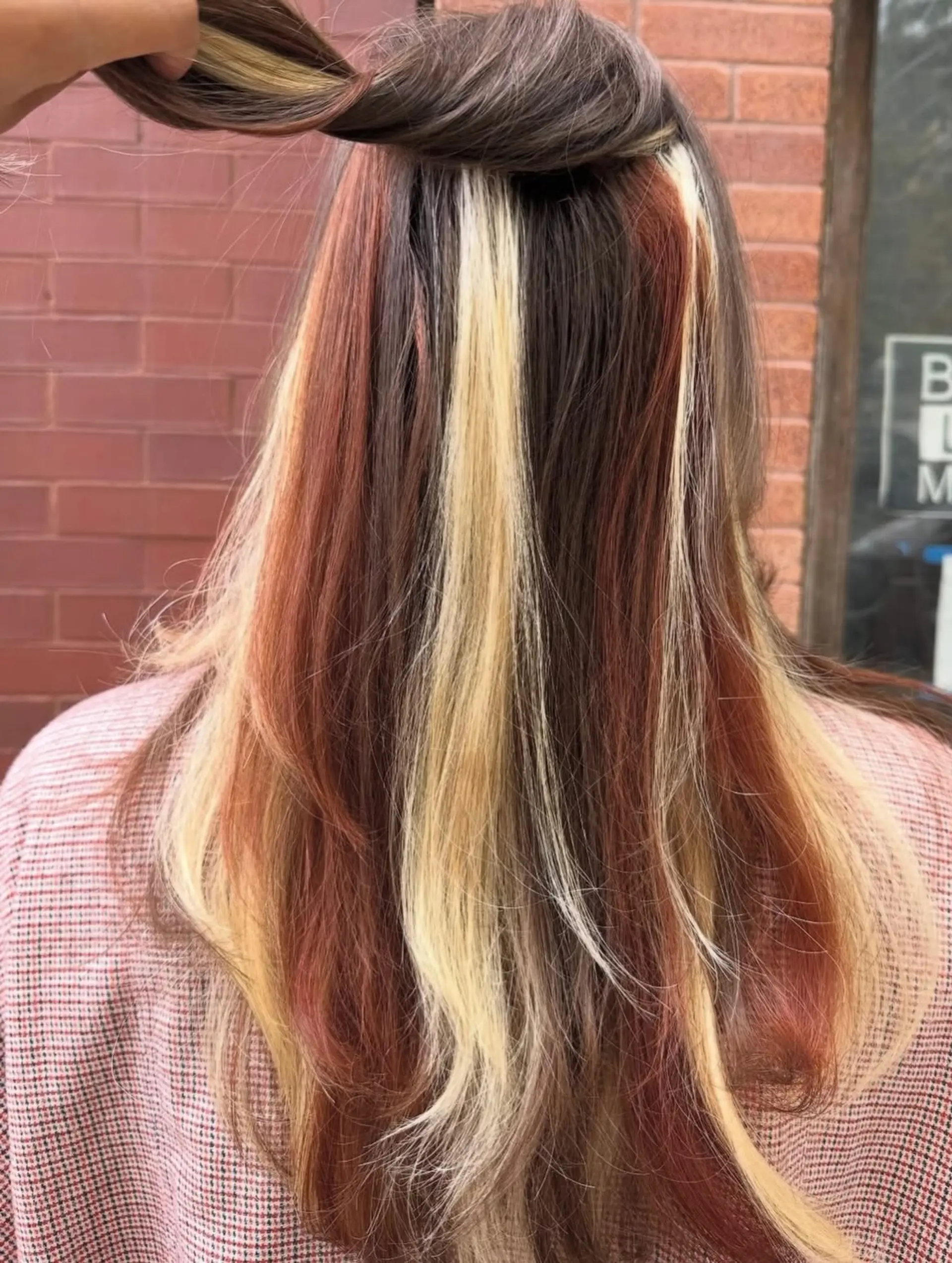 Long brown hair styled straight to showcase vibrant peekaboo color panels in blonde and auburn tones placed beneath the surface. 1