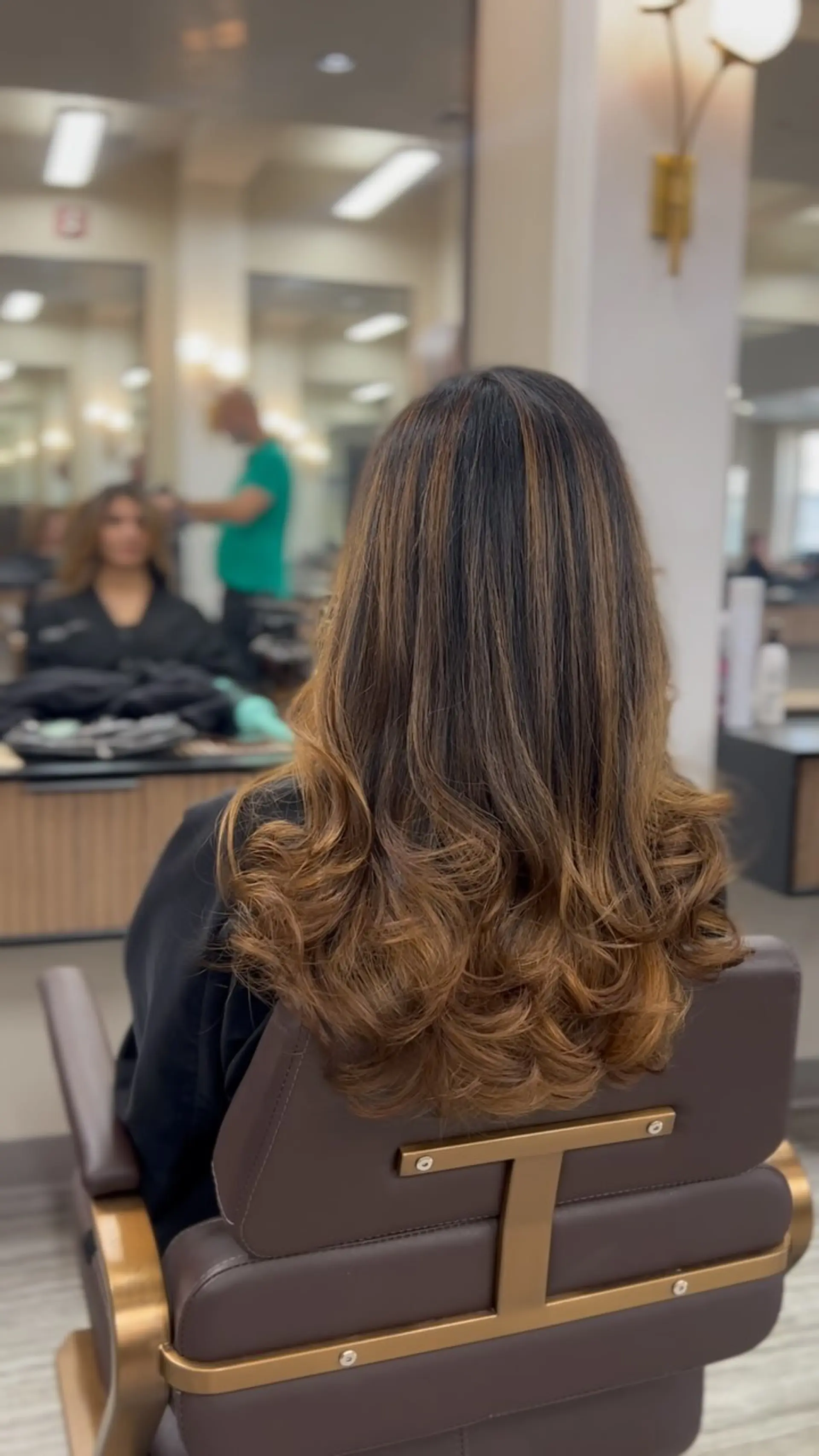 Long brunette hair enhanced with seamless warm caramel balayage highlights, styled in a voluminous blowout with bouncy curled ends. 1
