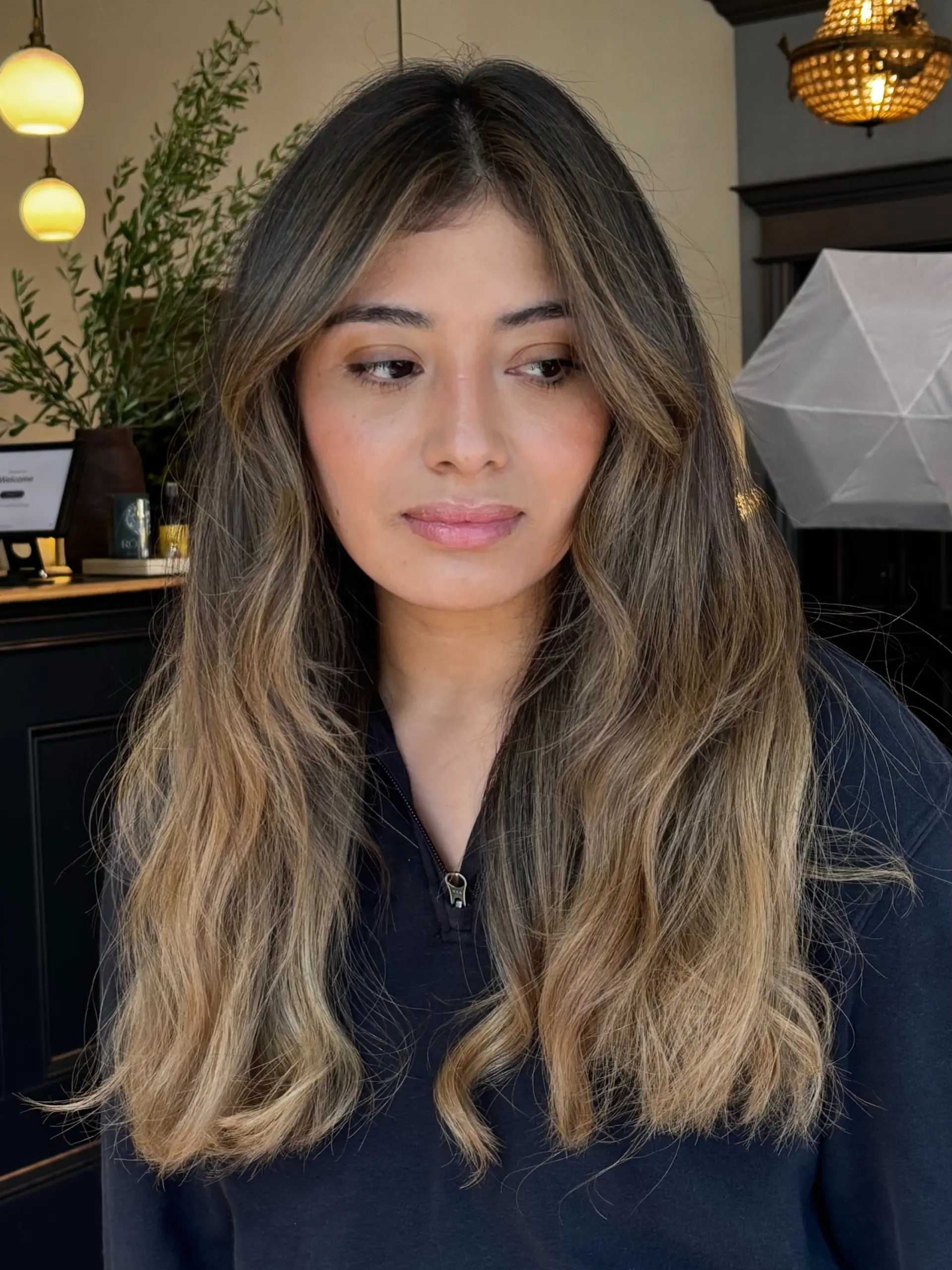 Long dimensional balayage featuring warm blonde and caramel ribbons blended into a dark brown base, styled with soft waves and curtain bangs. 1